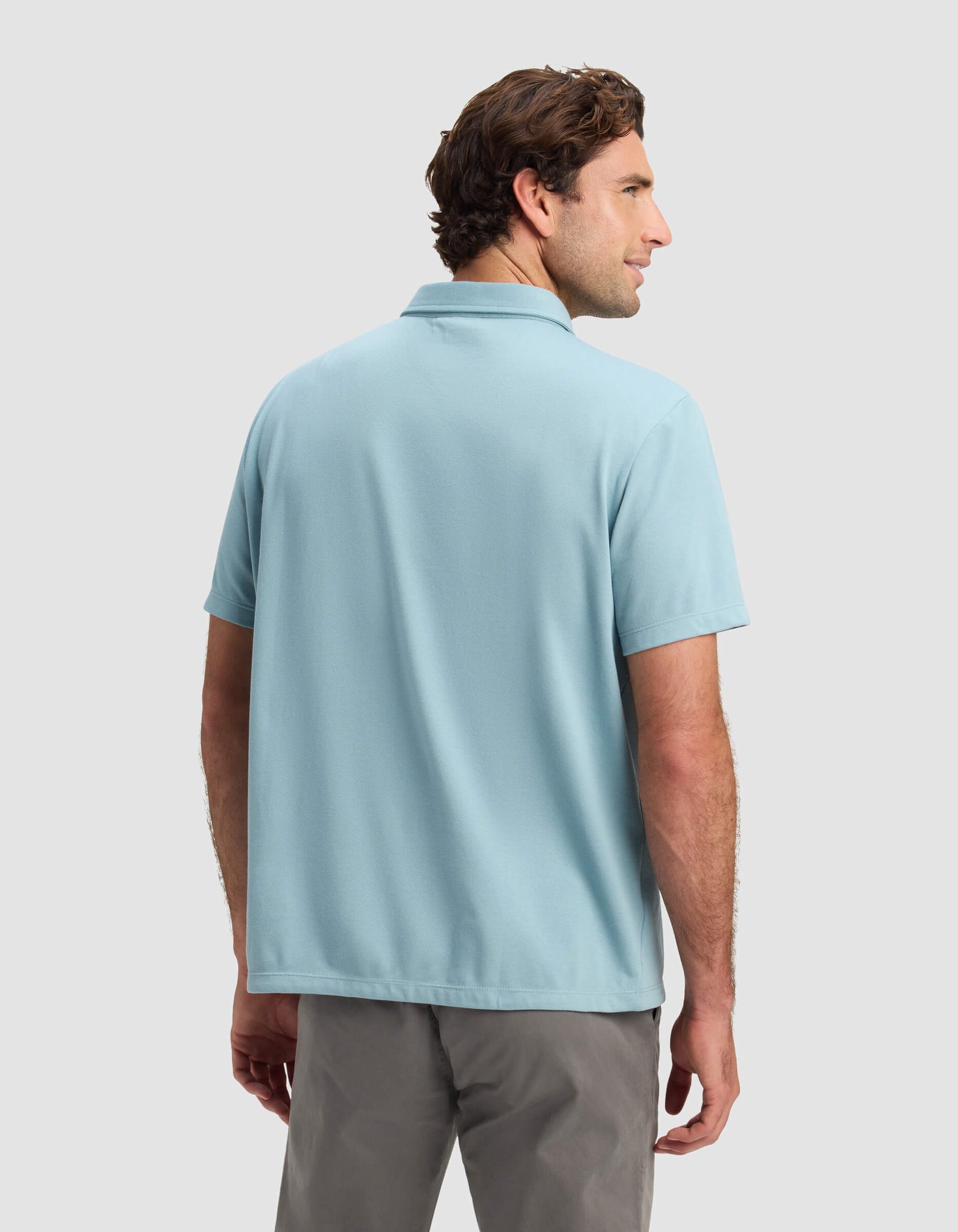 A man with short brown hair faces away, wearing a Cozy Earth Men's Pique Polo in light blue and gray pants against a plain, light gray background.