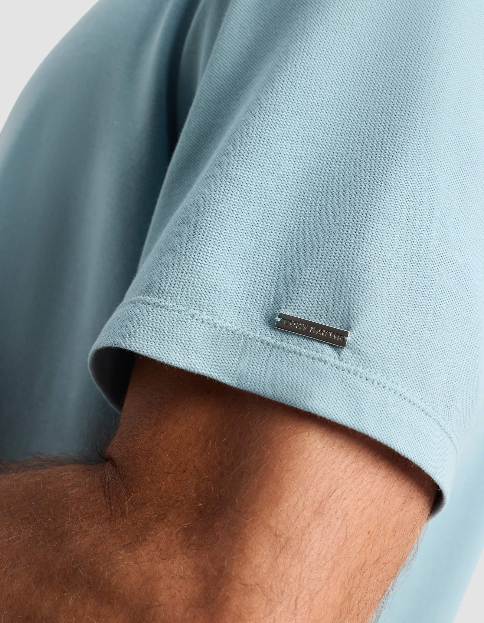 A close-up of a person’s arm wearing Cozy Earth’s Men’s Pique Polo in light blue, featuring a small metallic tag with engraved text near the sleeve hem.