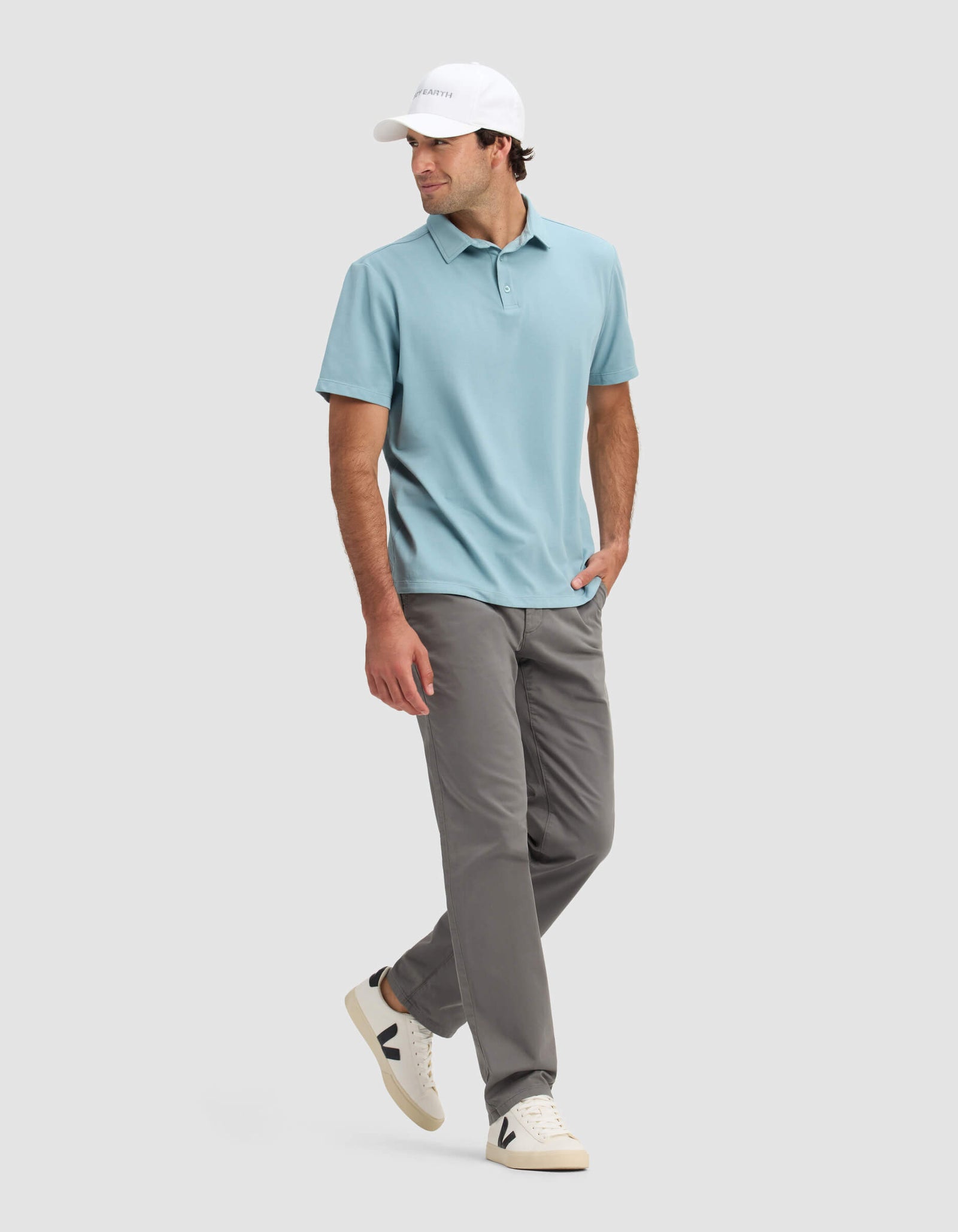 A man in Cozy Earth's Men's Pique Polo (light blue), gray pants, white sneakers with black accents, and a white cap stands against a light gray background with one hand in his pocket, looking to the side.