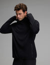 A man with wavy brown hair wears the Cozy Earth Men’s Recovery Hoodie in black, lifting its collar over his head. The background is plain gray, and the hoodie features a zippered pocket on the side.|Color:Black