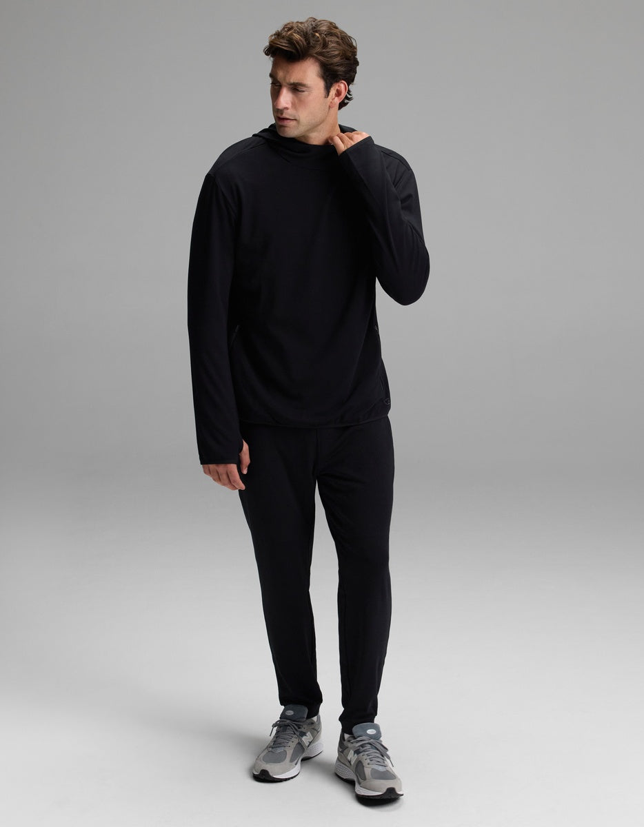 A man wearing the Cozy Earth Men’s Recovery Hoodie in black and matching black pants stands on a gray background, looking to his left with his right hand touching his neck. He completes the look with gray sneakers.