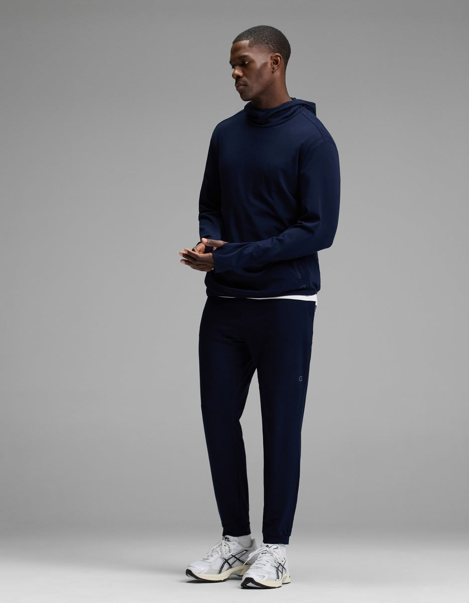 A man stands wearing the Cozy Earth Men’s Recovery Jogger in navy, paired with a matching navy hooded sweatshirt and white athletic sneakers, set against a plain gray background.|Color:Navy