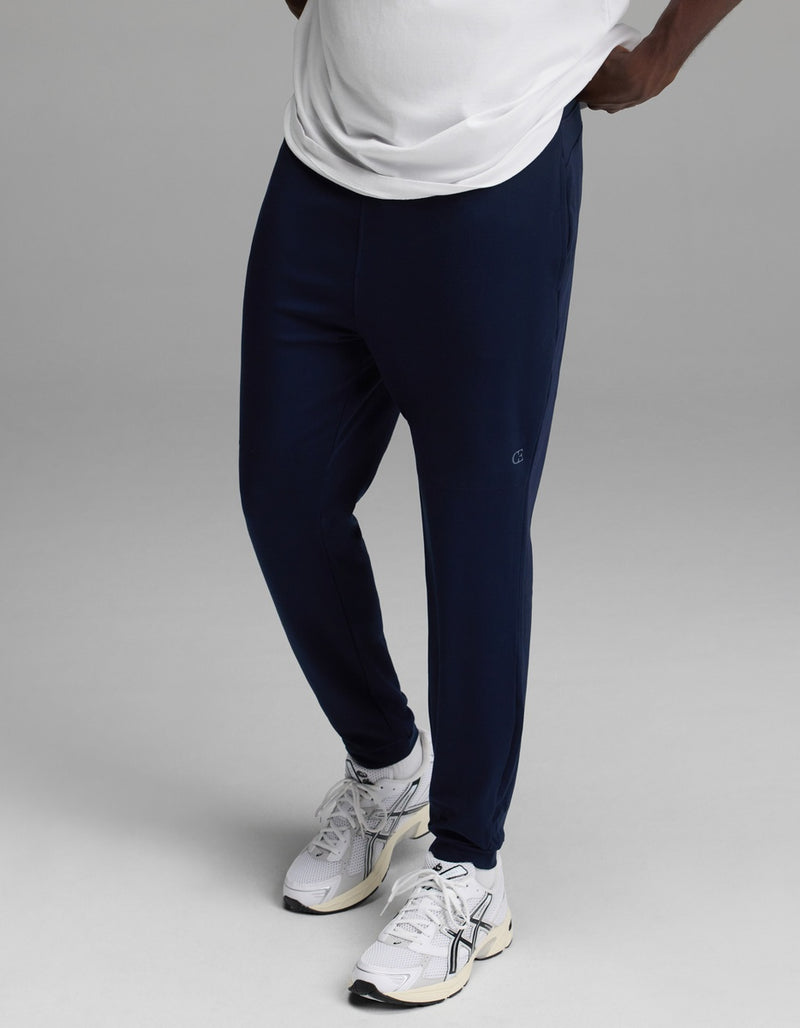 A person wearing a white shirt, Cozy Earth Men’s Recovery Jogger in navy, and white athletic sneakers stands against a plain gray background.