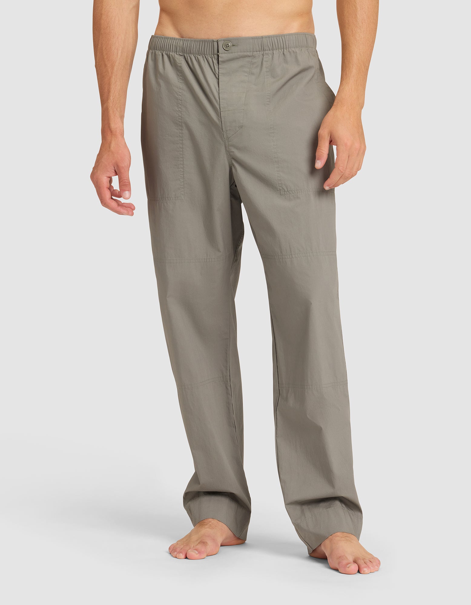 A man stands barefoot against a plain light background, wearing Cozy Earth Men's Sleep Pant in light gray with an elastic waistband and button closure. His torso is not visible. 