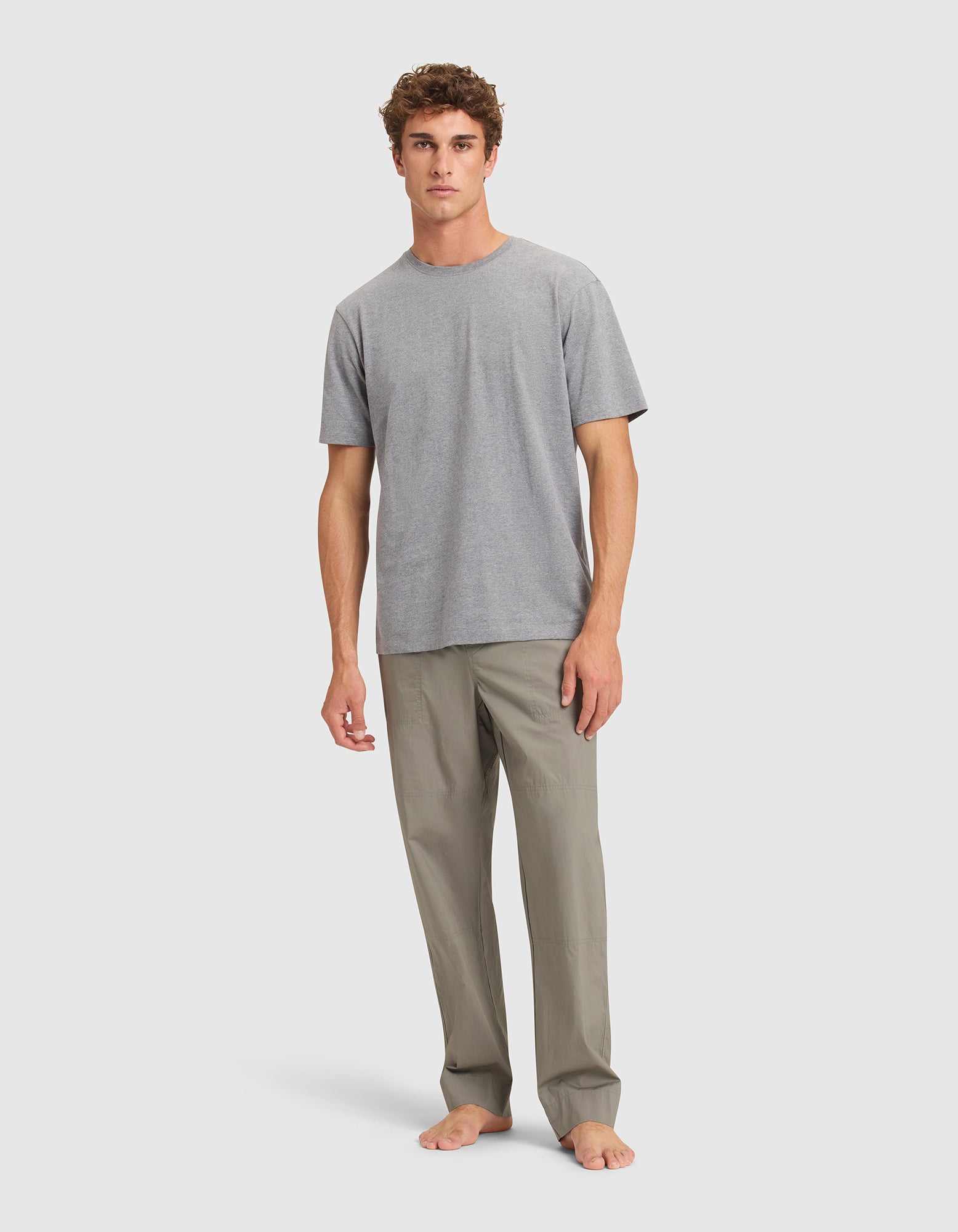 A man with short curly hair stands barefoot against a plain light background, wearing Cozy Earth Men's Sleep Pant and a plain grey T-shirt. |Color:Cypress