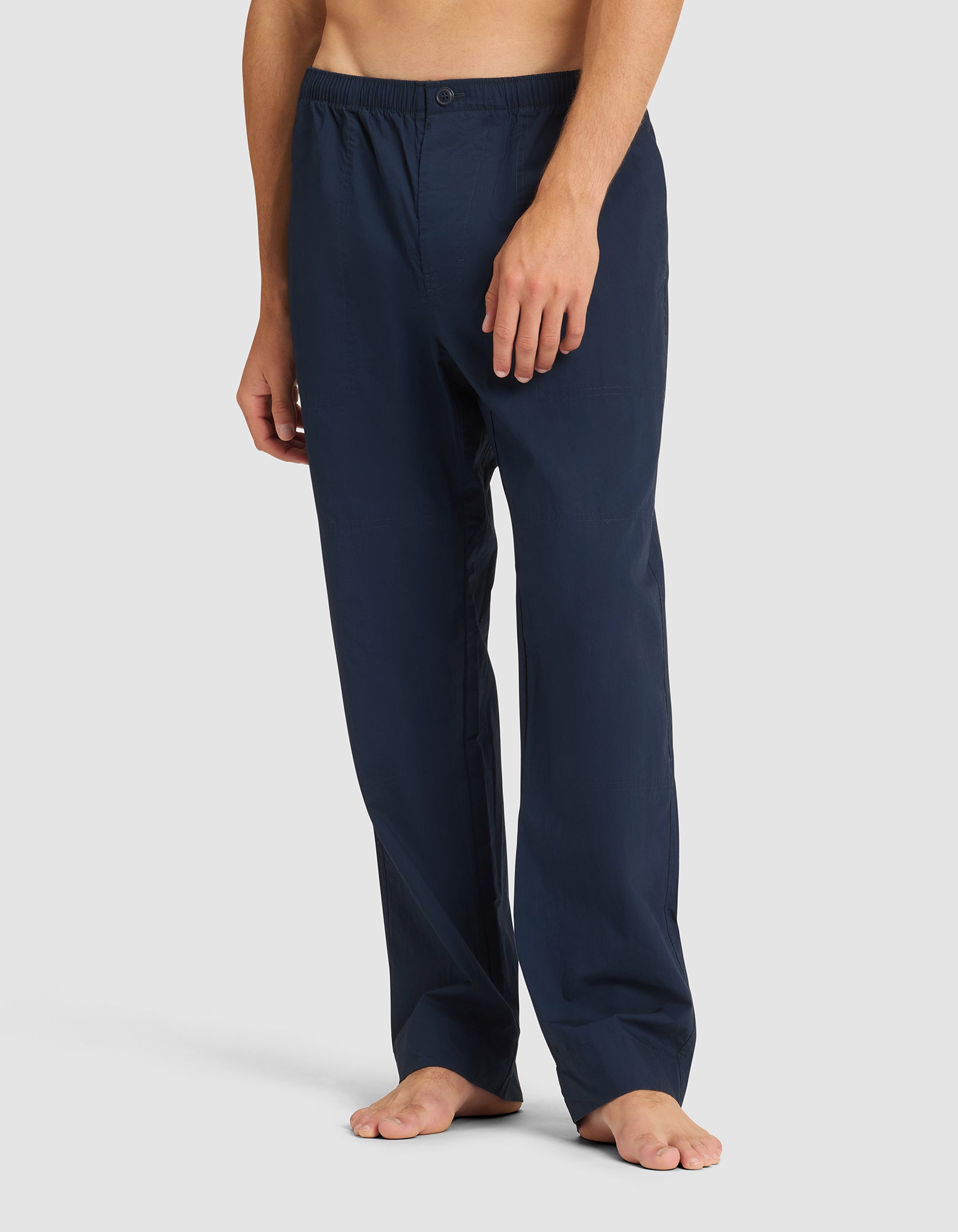 A person is wearing Cozy Earth Men's Sleep Pant in navy blue with an elastic waistband and button, standing barefoot against a plain light background. The upper body is not visible. 