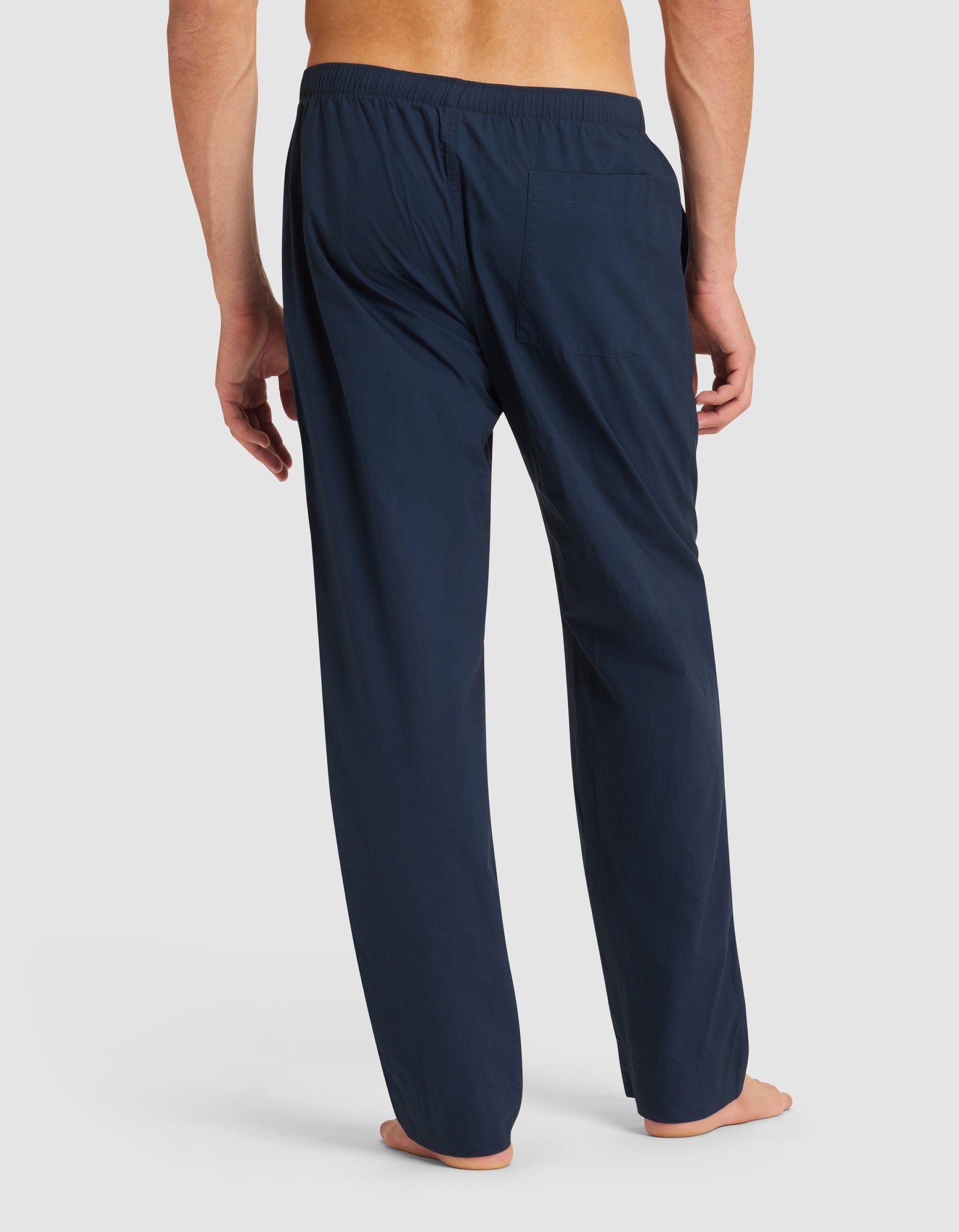 A person stands barefoot, shown from the back, wearing Cozy Earth Men's Sleep Pant—loose-fitting navy blue pajama pants with an elastic waistband and rear pocket—against a plain white background. 