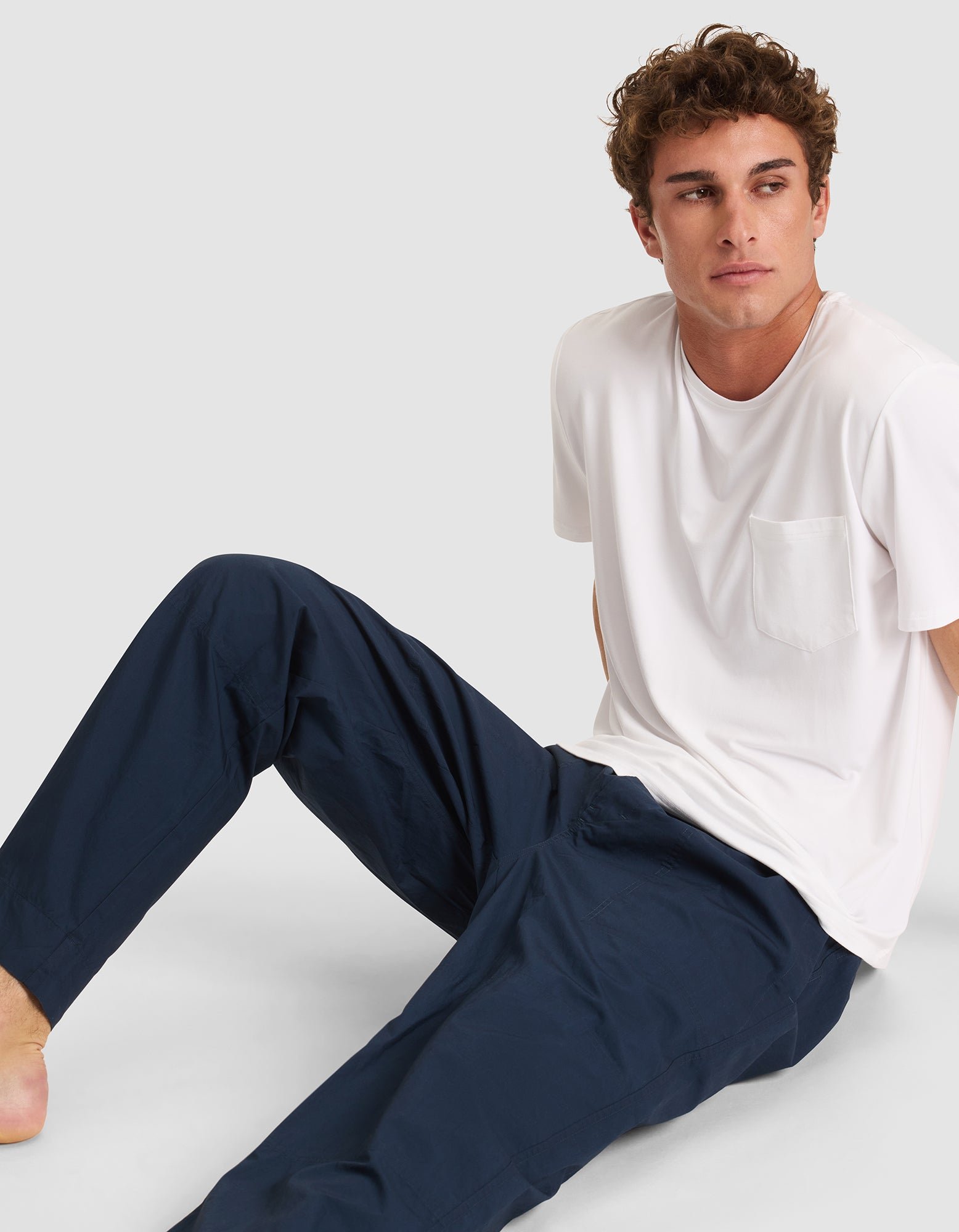 A young man with curly hair sits on the ground, wearing dark blue Cozy Earth Men's Sleep Pants and a white T-shirt with a chest pocket, looking to his left against a plain light background. 