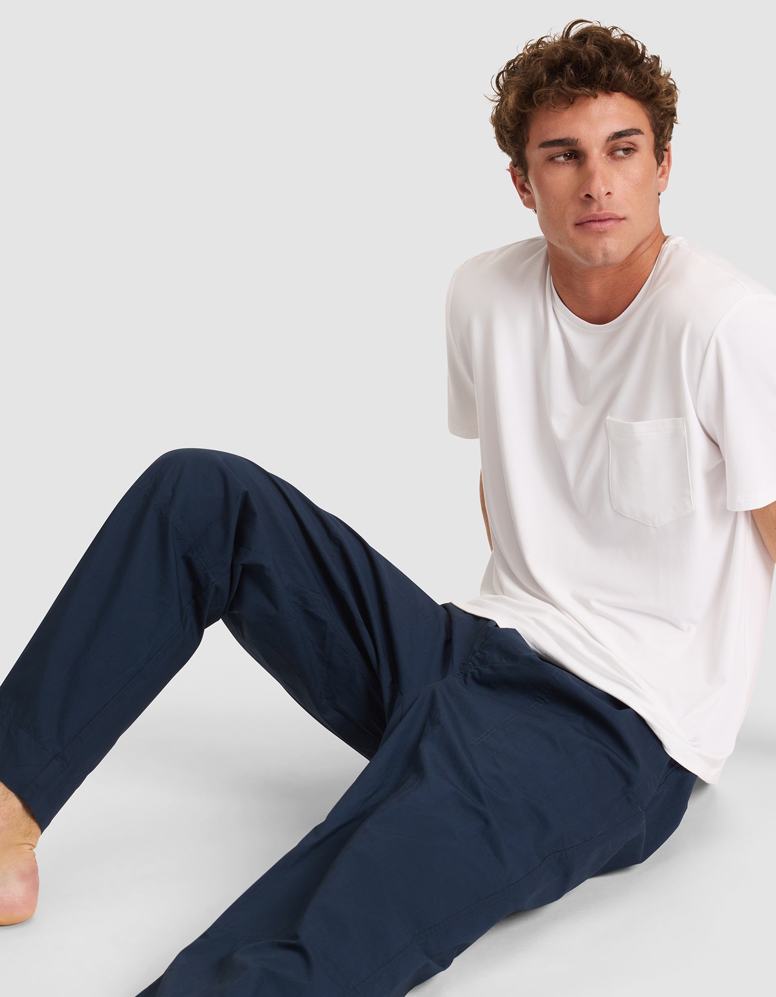 A young man with curly hair sits on the ground, wearing dark blue Cozy Earth Men's Sleep Pants and a white T-shirt with a chest pocket, looking to his left against a plain light background. 