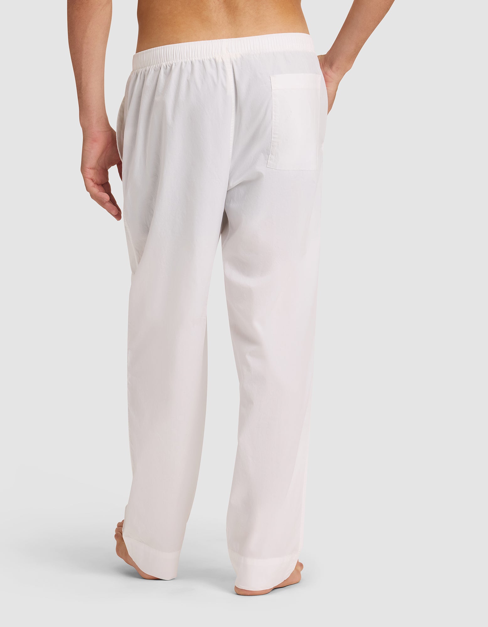 A person stands barefoot, seen from behind, wearing Cozy Earth Men's Sleep Pant—loose white pajama pants with an elastic waistband and right-side pocket—against a plain, light-colored background. 