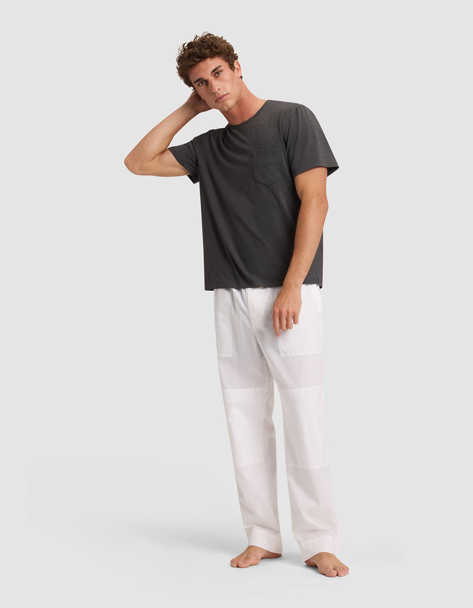 A man stands barefoot against a light background, looking relaxed with one hand on his head, dressed in Cozy Earth Men's Sleep Pant—loose white pants paired with a dark gray t-shirt. 