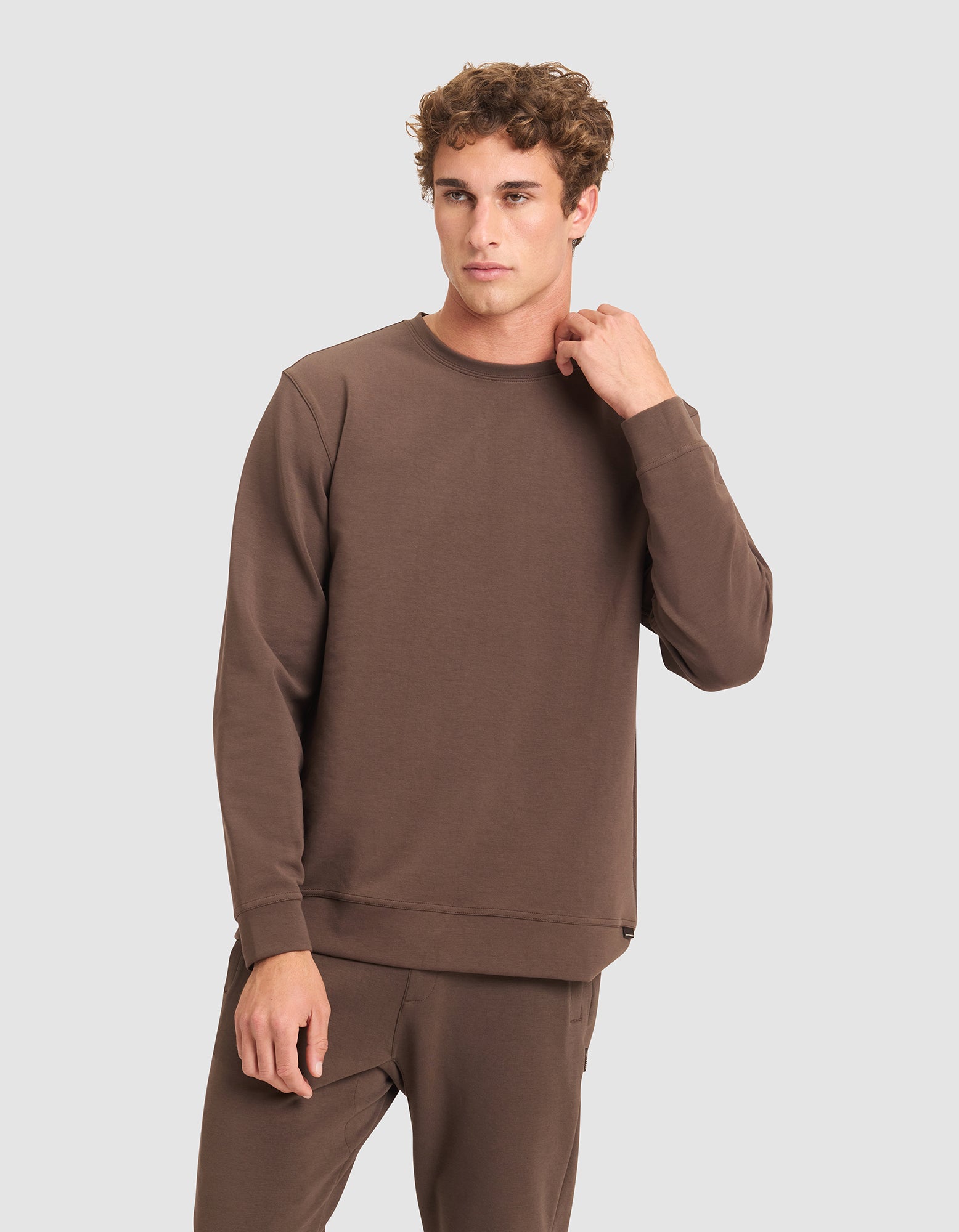 A man in Cozy Earth's Men's StretchTech Crewneck and matching pants stands against a plain light gray background, gazing to the side with one hand touching his neck. |Color:Cacao