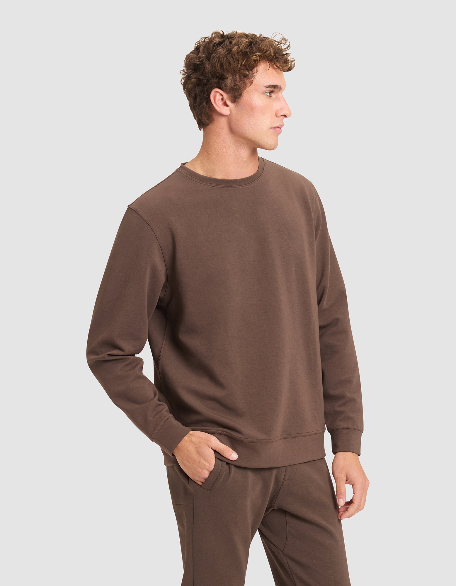 A young man with curly hair wears the Cozy Earth Men's StretchTech Crewneck and matching brown pants, standing with one hand in his pocket against a plain light gray background. 