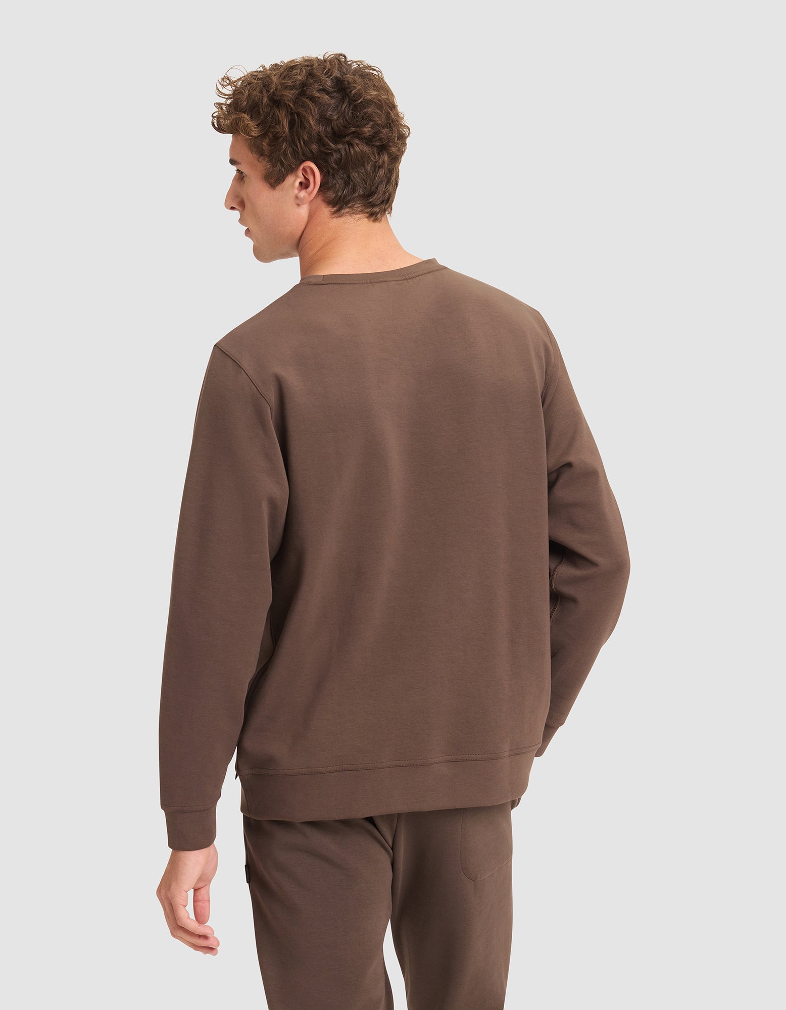 A man with curly hair stands facing away from the camera, wearing Cozy Earth’s Men's StretchTech Crewneck in brown with matching pants, against a light gray background. 