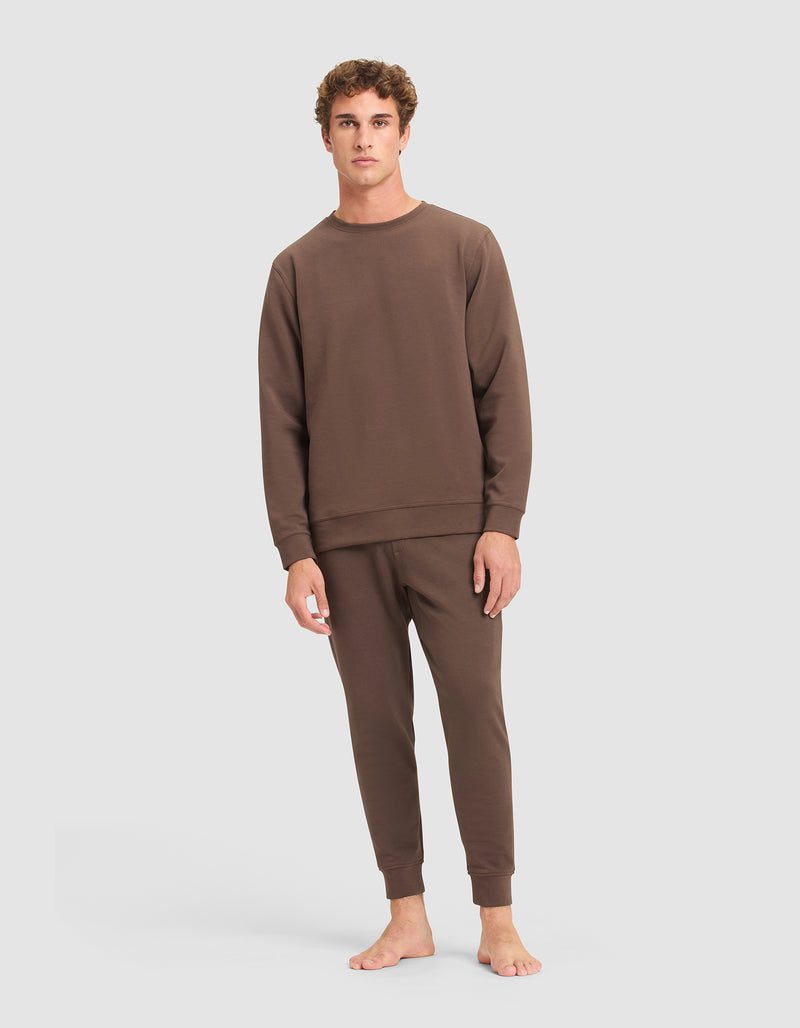 A young man with short curly hair stands barefoot against a plain background, wearing Cozy Earth’s Men's StretchTech Crewneck and matching joggers, facing forward with a neutral expression and relaxed posture.