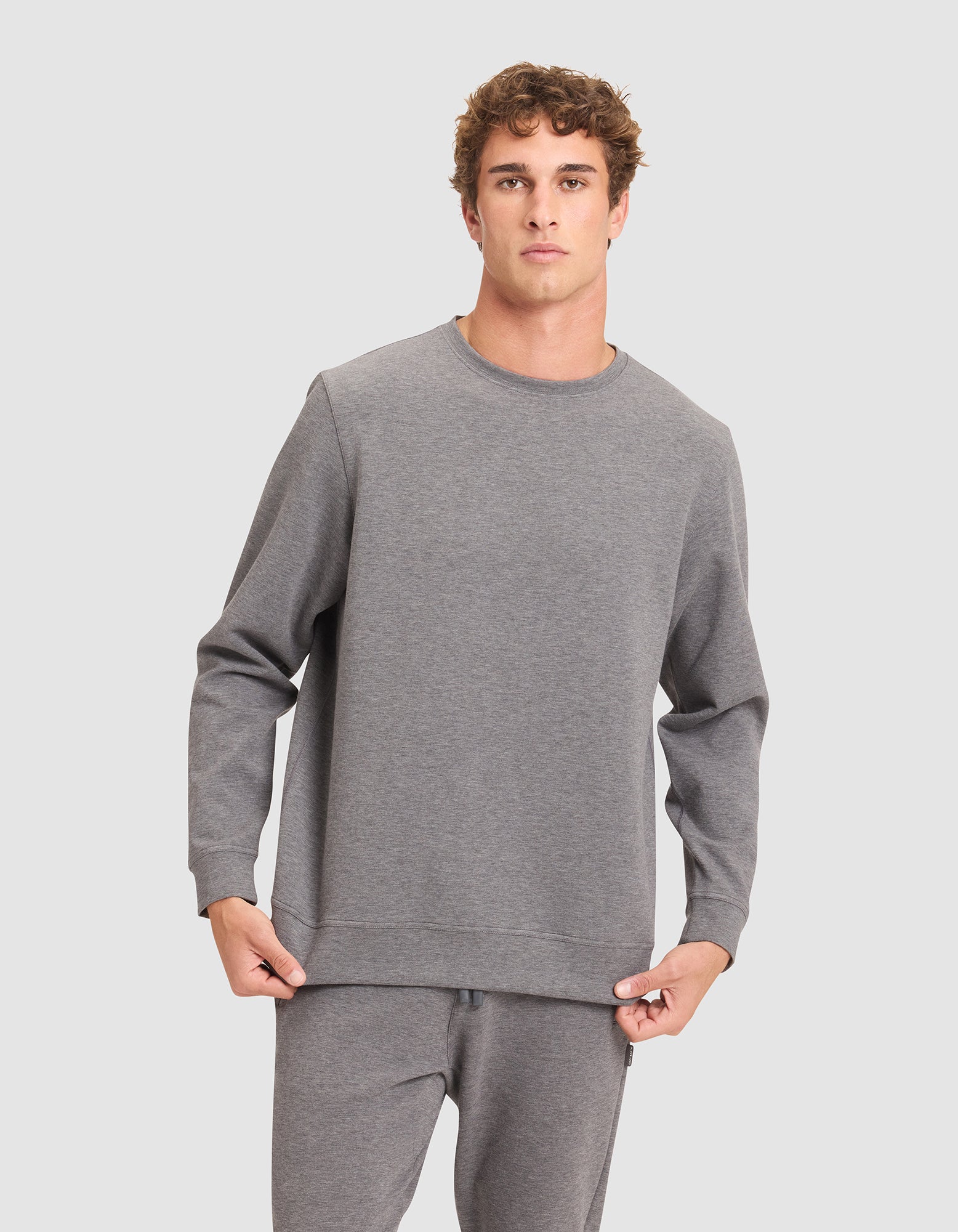 A young man in a Cozy Earth Men's StretchTech Crewneck and matching sweatpants stands before a plain light gray background, looking at the camera while holding the hem of his sweatshirt. 