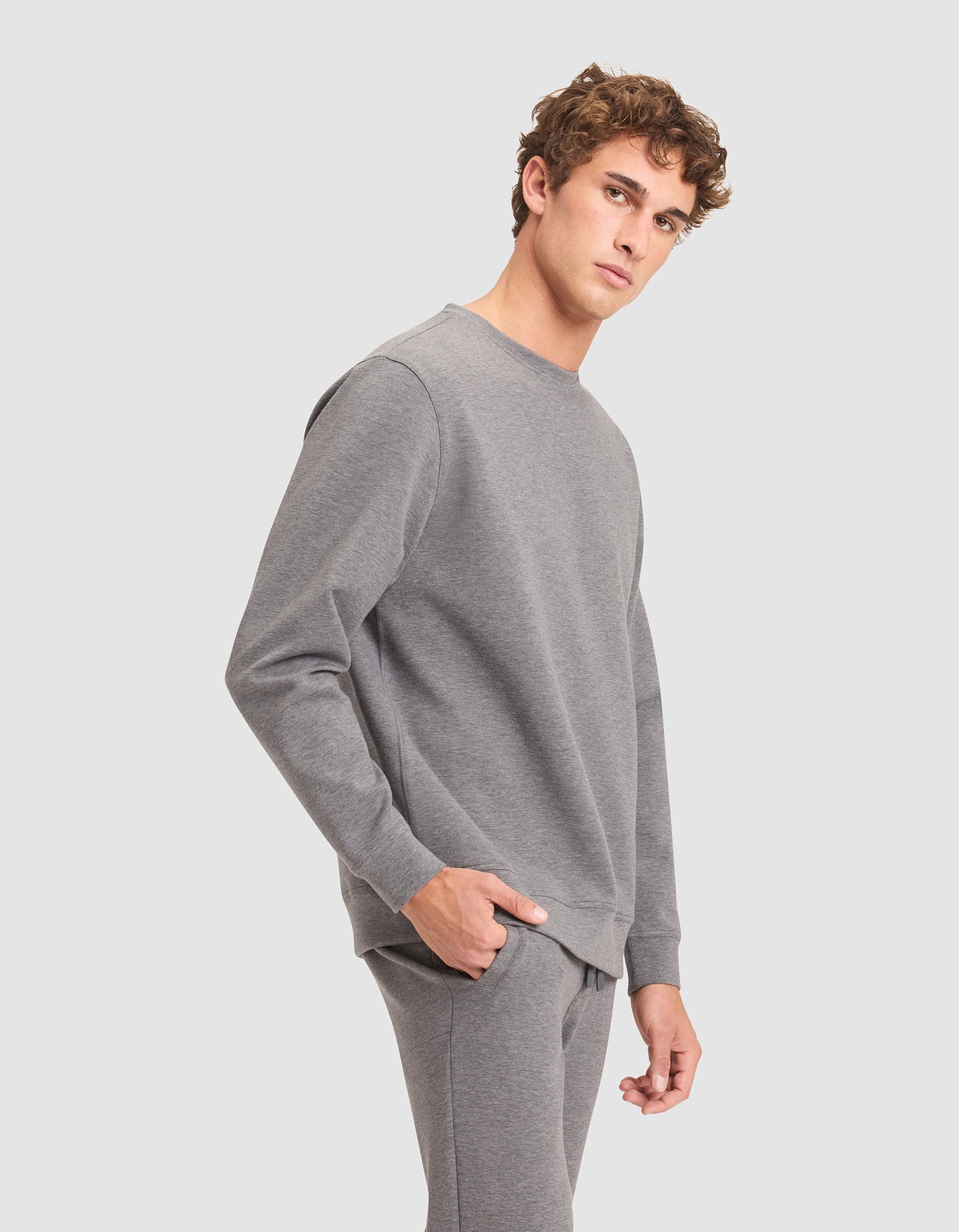 A young man with curly hair, wearing the Cozy Earth Men's StretchTech Crewneck and matching sweatpants, stands with one hand in his pocket, gazing to the side against a plain light grey background. 