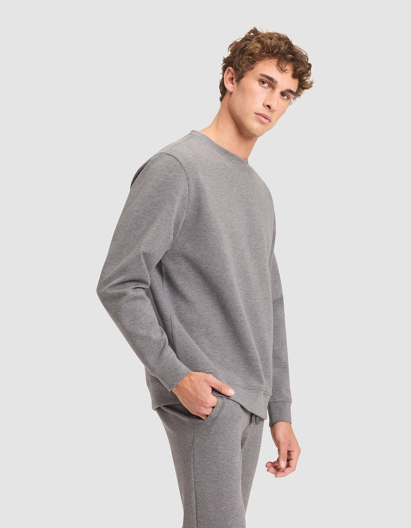 A young man with curly hair, wearing the Cozy Earth Men's StretchTech Crewneck and matching sweatpants, stands with one hand in his pocket, gazing to the side against a plain light grey background.