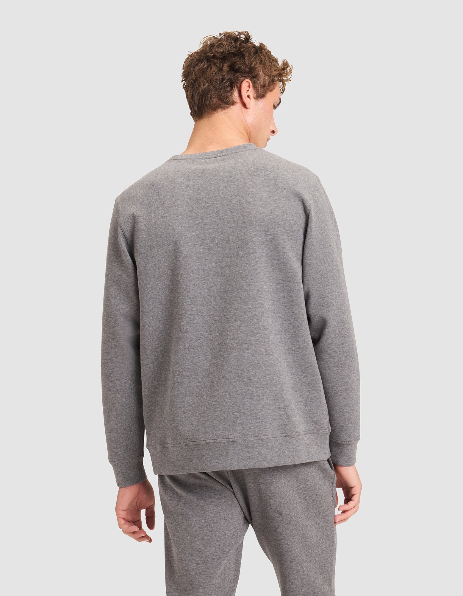 A man with short curly hair wears the Cozy Earth Men's StretchTech Crewneck in grey, paired with matching sweatpants. He faces away from the camera, glancing over his left shoulder against a plain light grey background. 