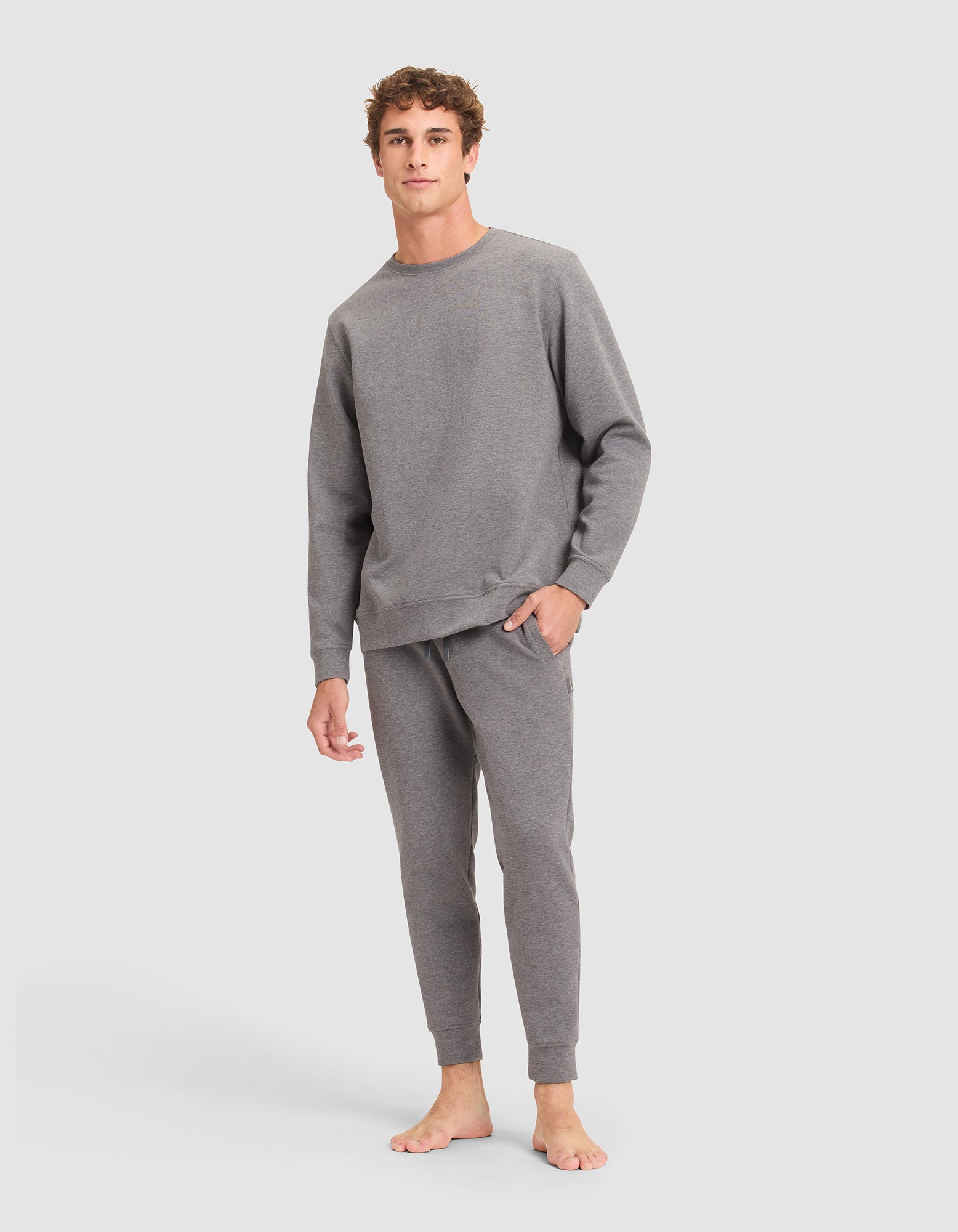 A young man stands barefoot against a light gray background, wearing Cozy Earth's Men's StretchTech Crewneck and matching jogger pants. He has short curly hair, a relaxed expression, and one hand in his pocket. 