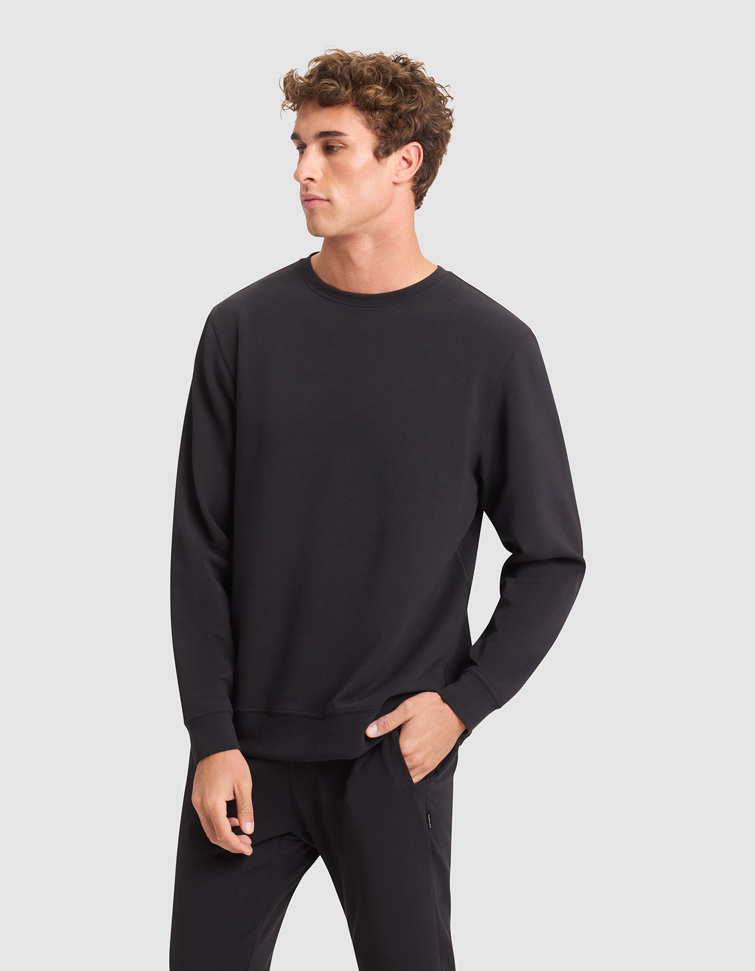 A young man with curly brown hair models the Cozy Earth Men's StretchTech Crewneck and matching black pants, standing against a light gray background with one hand in his pocket, gazing to the side. 