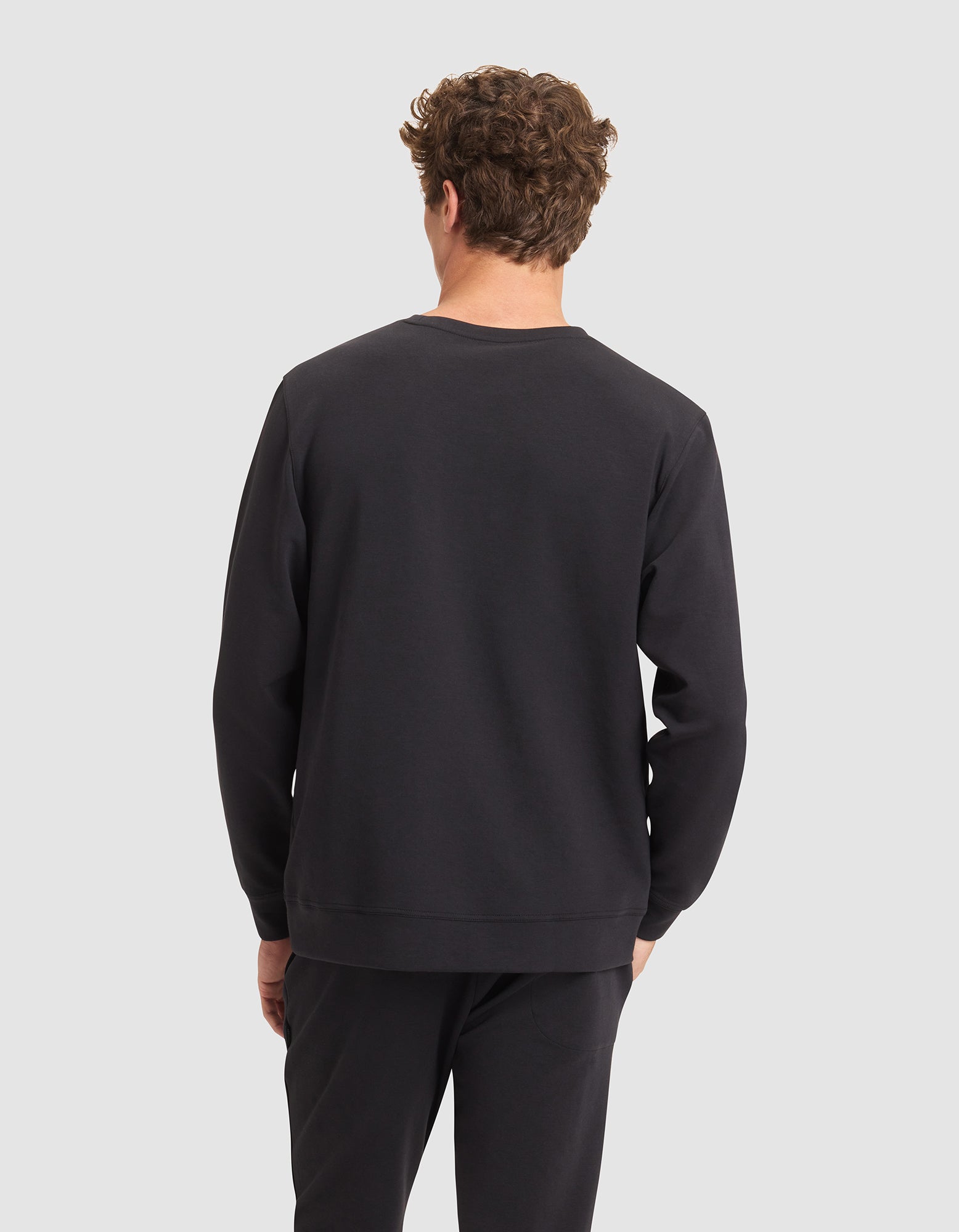 A person with short curly hair stands facing away from the camera, wearing a Cozy Earth Men's StretchTech Crewneck and black pants against a light gray background. 