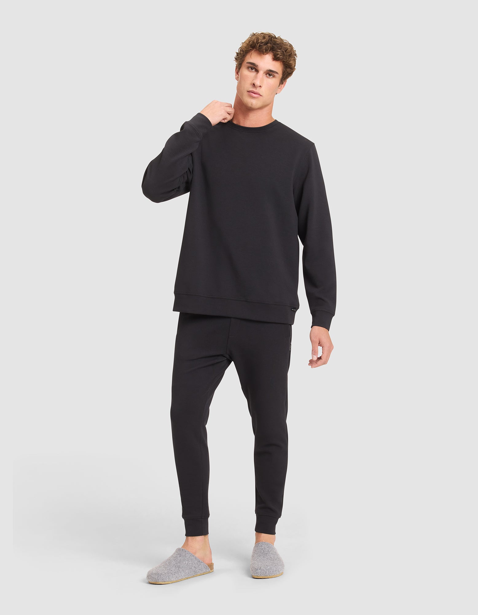 A man with curly brown hair stands against a light background wearing Cozy Earth’s Men's StretchTech Crewneck in black. He matches it with black joggers and gray slippers, looking at the camera with a neutral expression. 