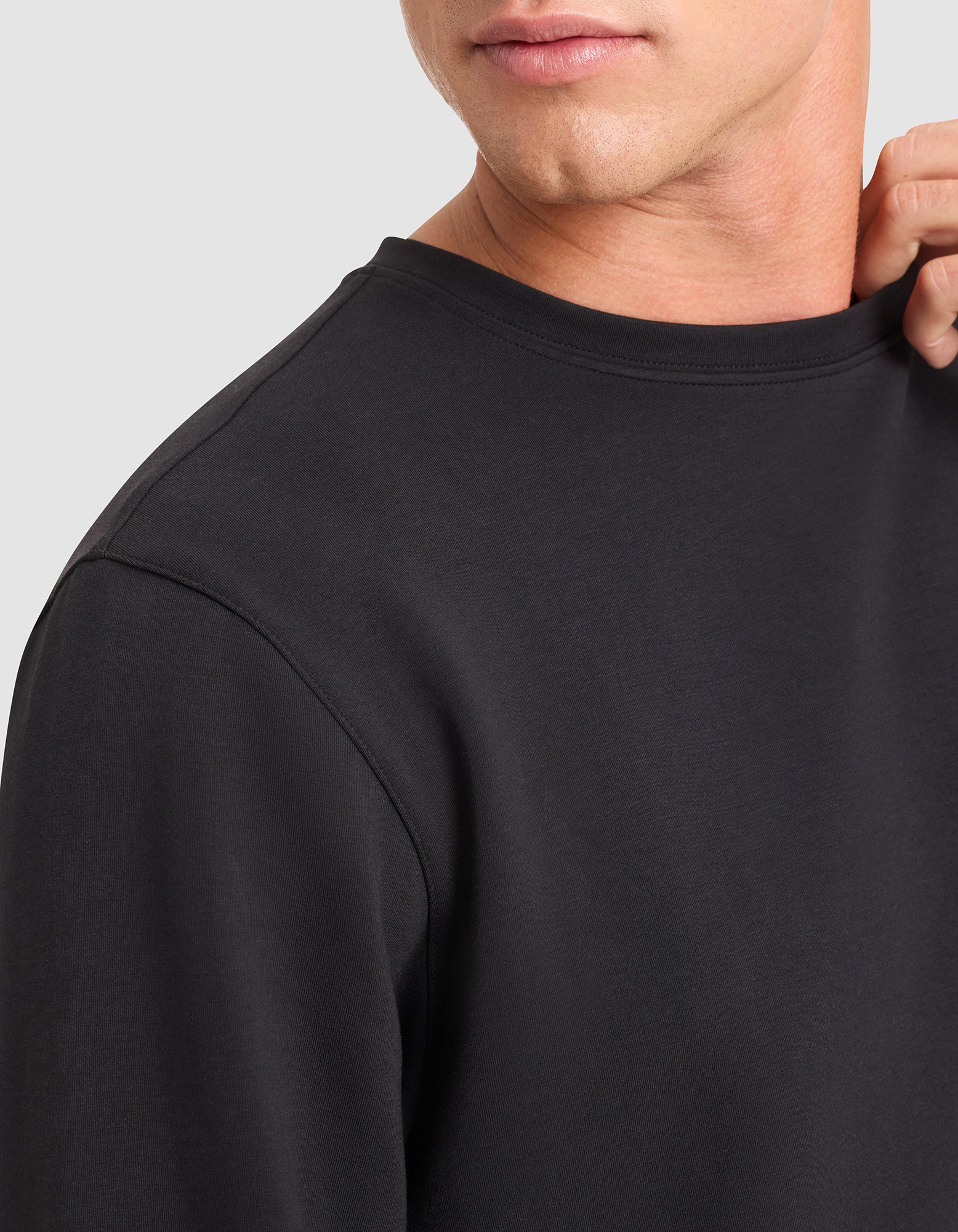 A close-up of a person wearing the Cozy Earth Men's StretchTech Crewneck in black, gently holding the collar. The image displays part of their face, neck, and upper torso. 
