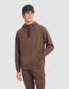 A man wears the Cozy Earth Men's StretchTech Hoodie in brown with a kangaroo pocket, paired with matching brown pants and a black baseball cap, standing against a plain light gray background.  |Color:Cacao
