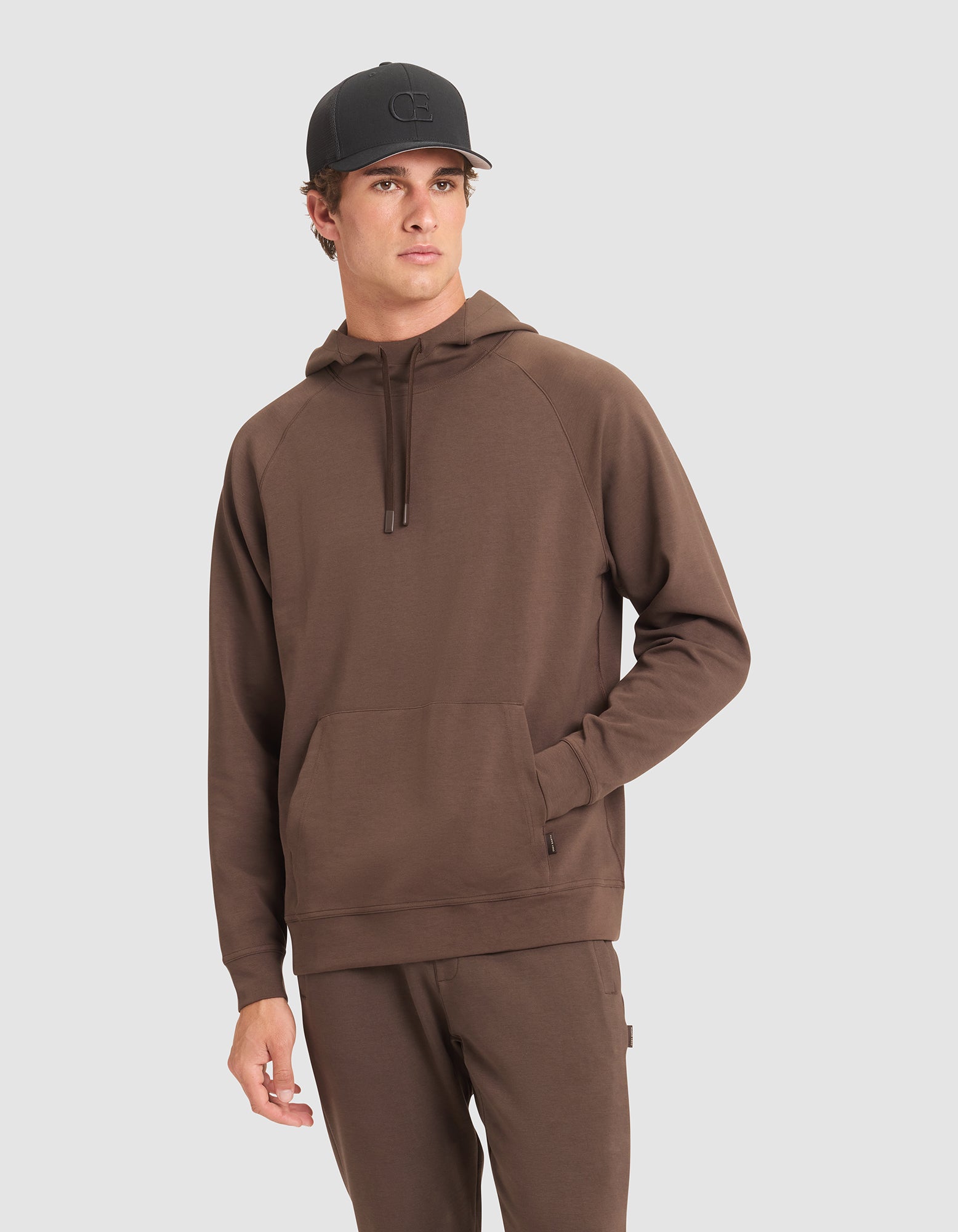 A man wears the Cozy Earth Men's StretchTech Hoodie in brown with a kangaroo pocket, paired with matching brown pants and a black baseball cap, standing against a plain light gray background.  