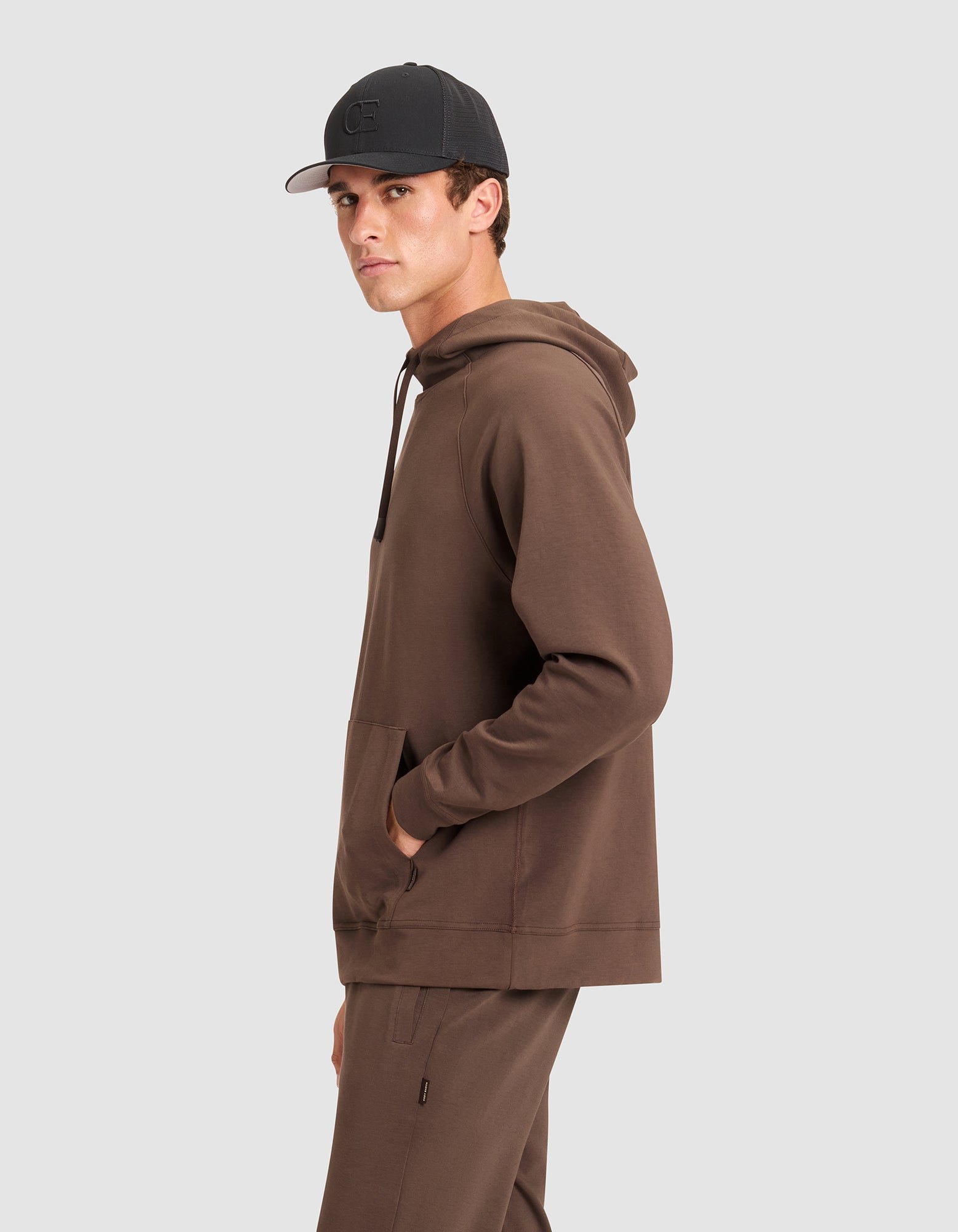 A young man in profile wears a Cozy Earth Men's StretchTech Hoodie and matching pants with one hand in his pocket, paired with a black baseball cap. He glances toward the camera against a plain light gray background.
