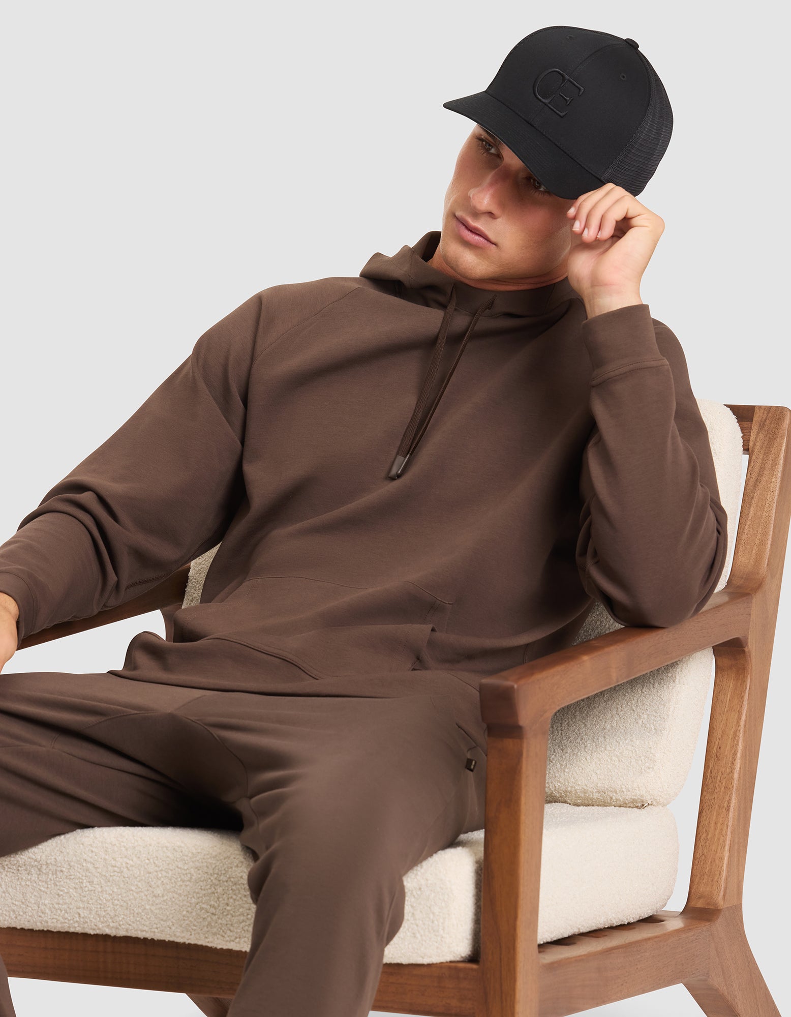 A man wearing the Cozy Earth Men's StretchTech Hoodie in brown and matching pants sits on a wooden chair with cream cushions, resting his head on one hand and wearing a black baseball cap.