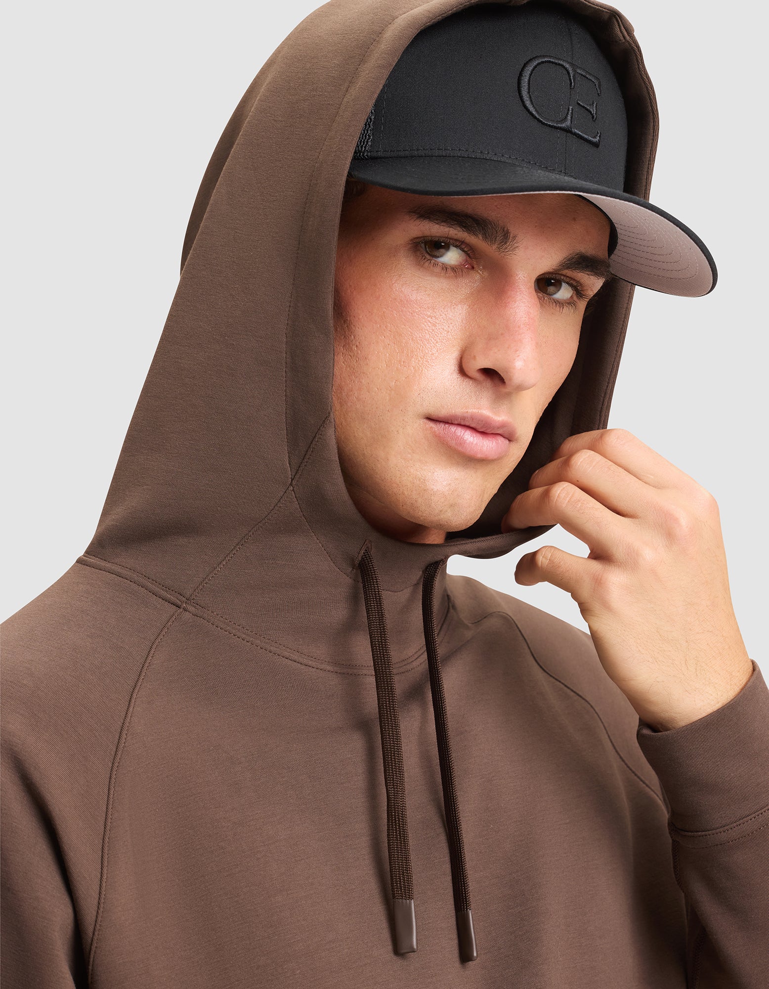 A man in a Cozy Earth Men's StretchTech Hoodie and a black cap with embroidered letters looks at the camera, holding the edge of his hood.  