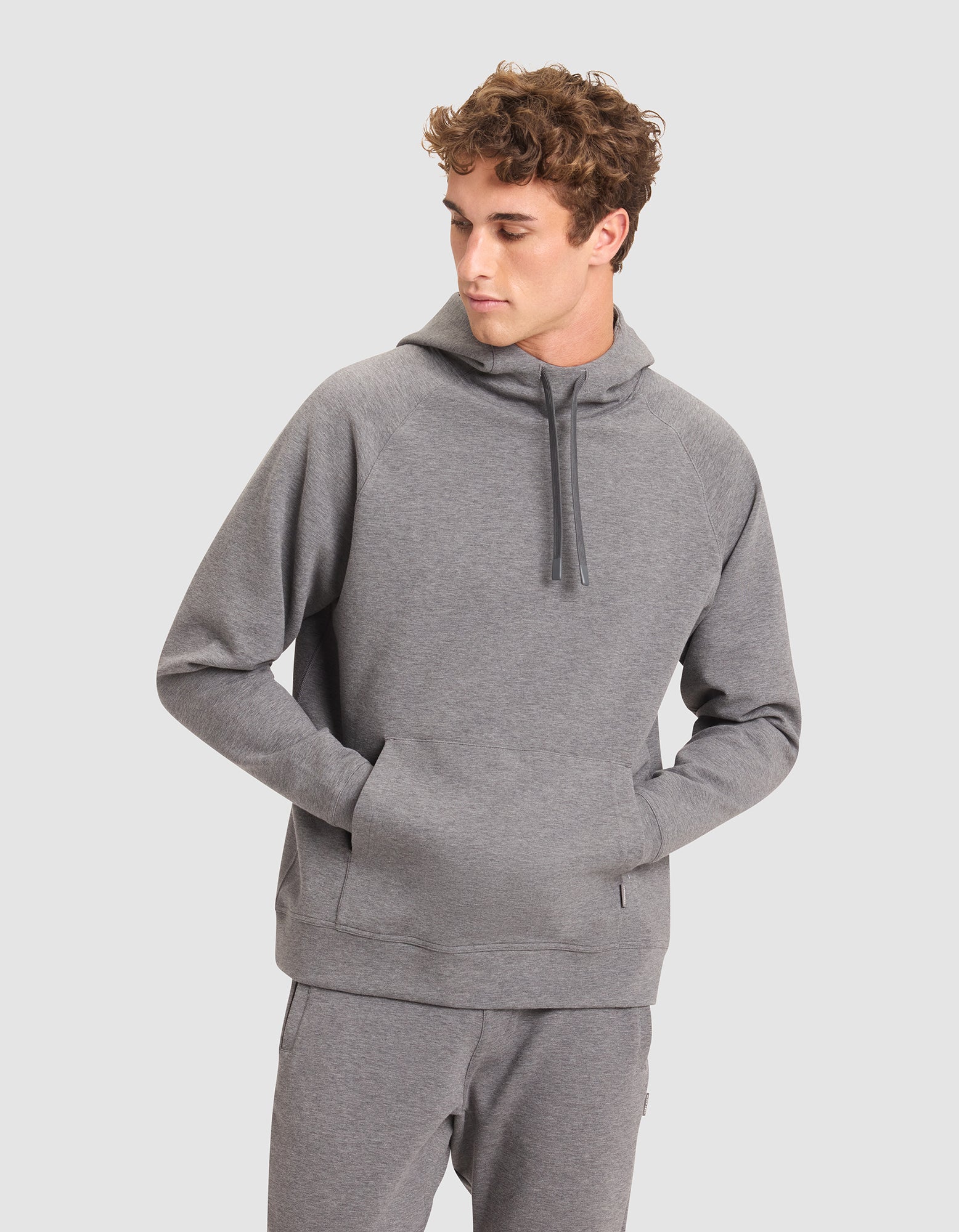 A young man wears the Cozy Earth Men's StretchTech Hoodie in light gray with matching sweatpants, hands in the hoodie pocket, looking left against a plain light gray background. |Color:Coal Heather