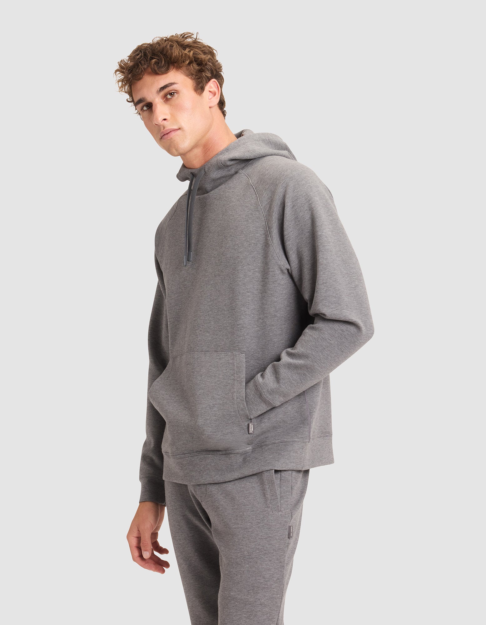 A young man models the Cozy Earth Men's StretchTech Hoodie in light grey, featuring a front pocket and relaxed fit sweatpants, standing against a plain backdrop and gazing slightly to the side. 