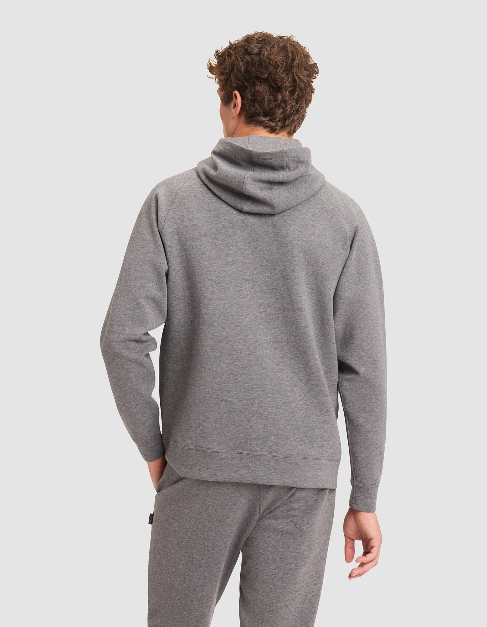 A man with curly hair is seen from behind wearing the Cozy Earth Men's StretchTech Hoodie in gray against a plain light background. 