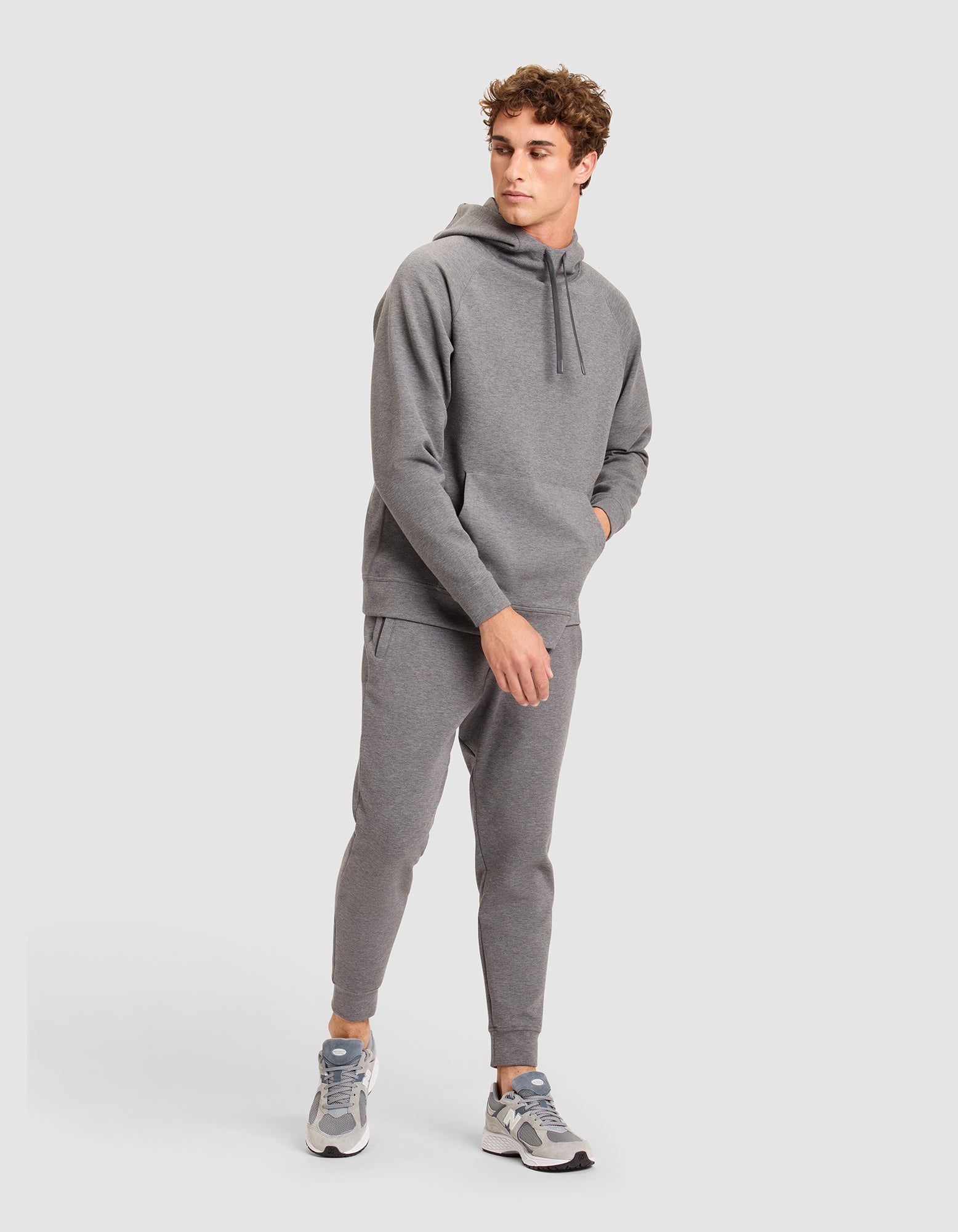 A man stands against a plain background wearing the Cozy Earth Men's StretchTech Hoodie and matching joggers, paired with gray and white sneakers. He has one hand in his pocket and looks to the side. 