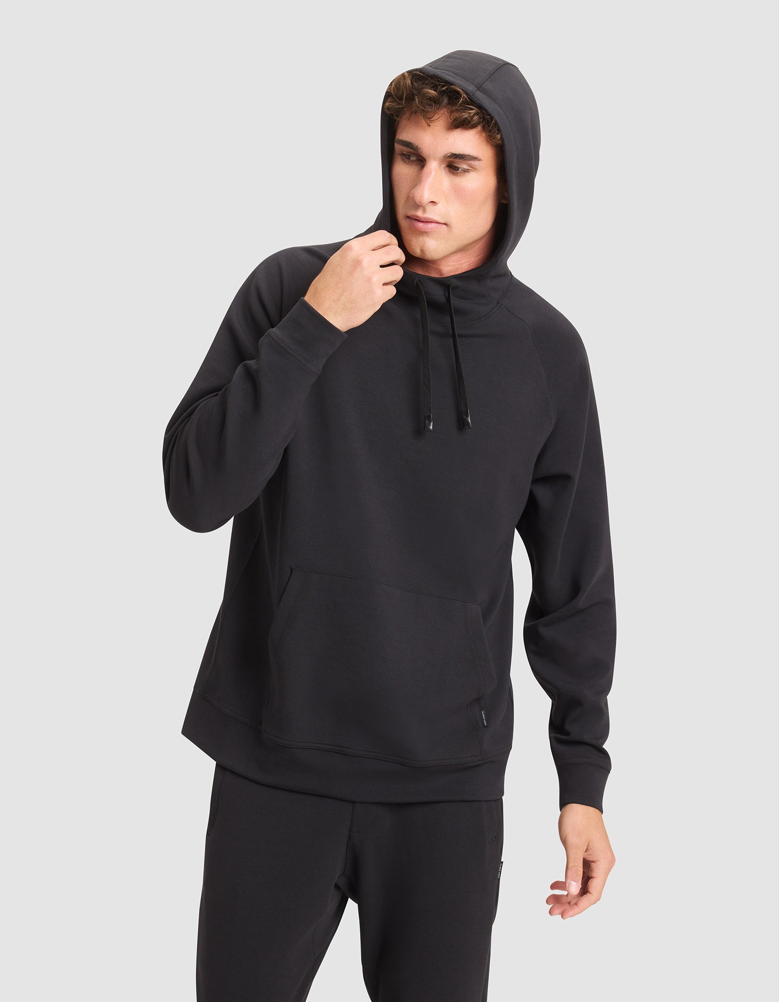 A young man wears the Cozy Earth Men's StretchTech Hoodie and black pants against a light gray background, looking to the side as he adjusts the hood. 