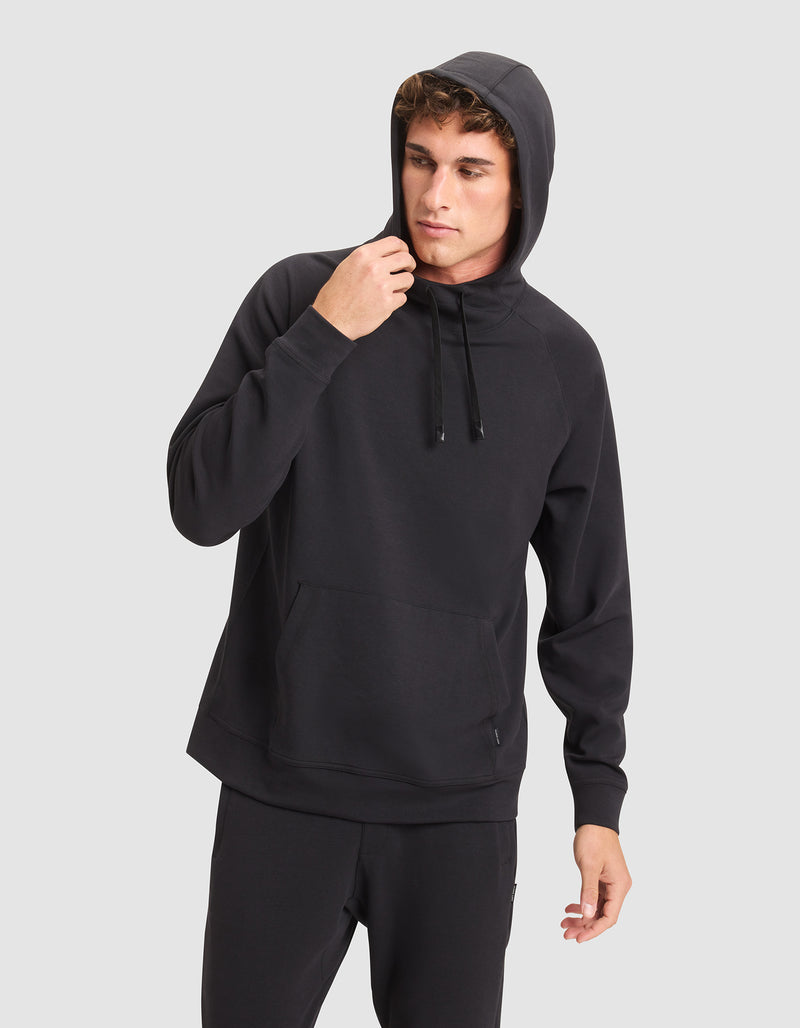 A young man wears the Cozy Earth Men's StretchTech Hoodie and black pants against a light gray background, looking to the side as he adjusts the hood.