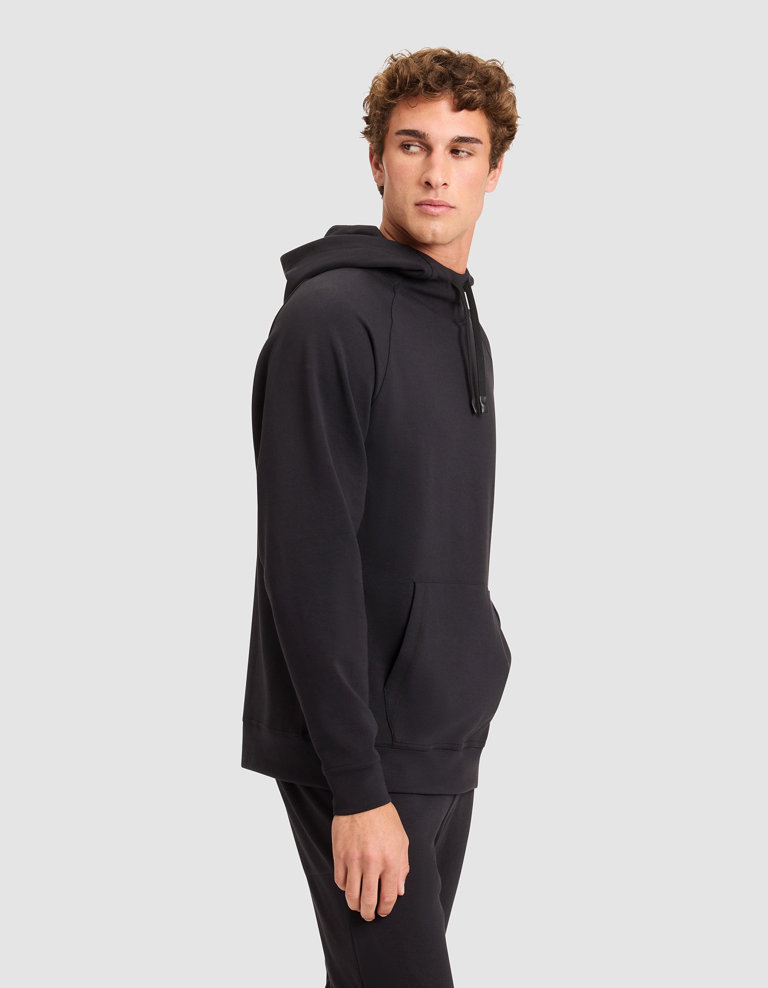 A young man wears the Cozy Earth Men's StretchTech Hoodie in black and matching pants, standing against a plain light gray background while looking to his left with his right hand at his side. 