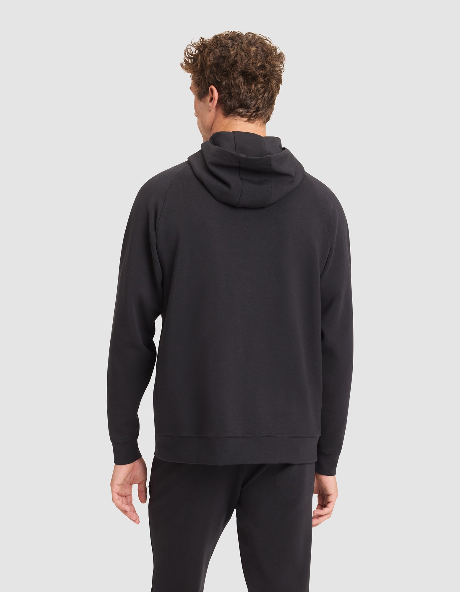A man with curly hair faces away from the camera, wearing a Cozy Earth Men's StretchTech Hoodie in black and black pants against a light gray background. 