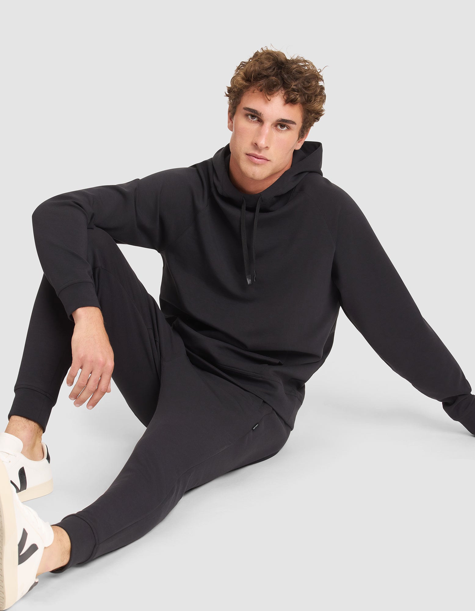 A young man with curly hair wears the Cozy Earth Men's StretchTech Hoodie and matching joggers, paired with white sneakers. He sits on the floor against a light gray background, looking at the camera with one knee up and an arm resting on his leg. 