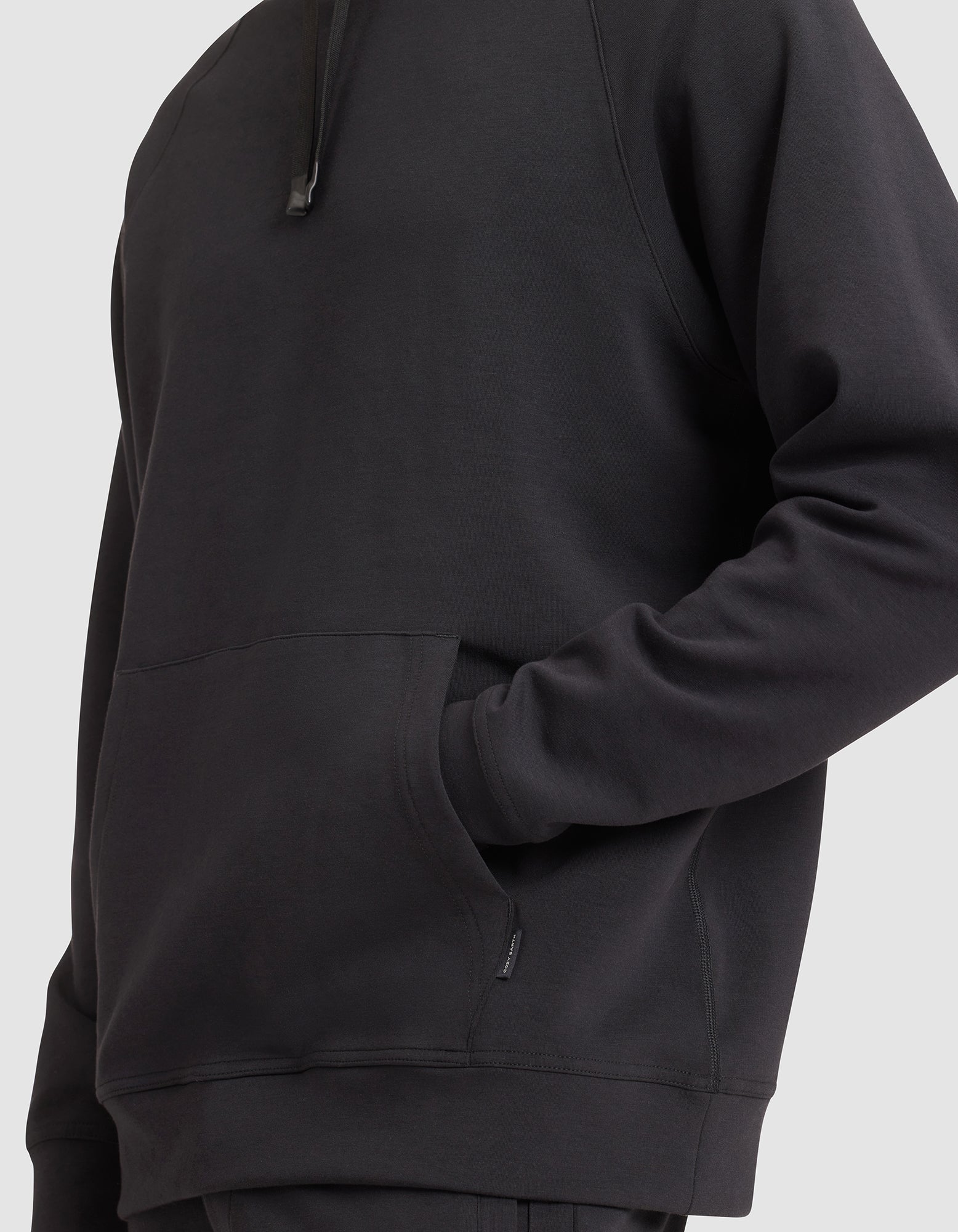 A man models the Cozy Earth Men's StretchTech Hoodie in black, one hand in the front pocket, shown from shoulders to hips against a plain light background. 