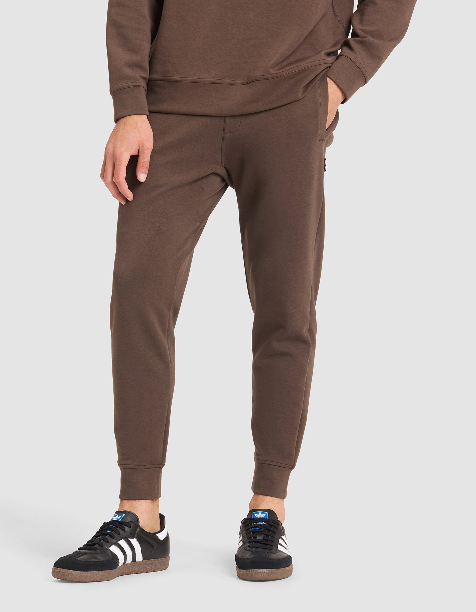 A person wears Cozy Earth Men's StretchTech Jogger sweatpants with a matching brown sweatshirt and black Adidas sneakers with white stripes, standing against a plain light gray background. 