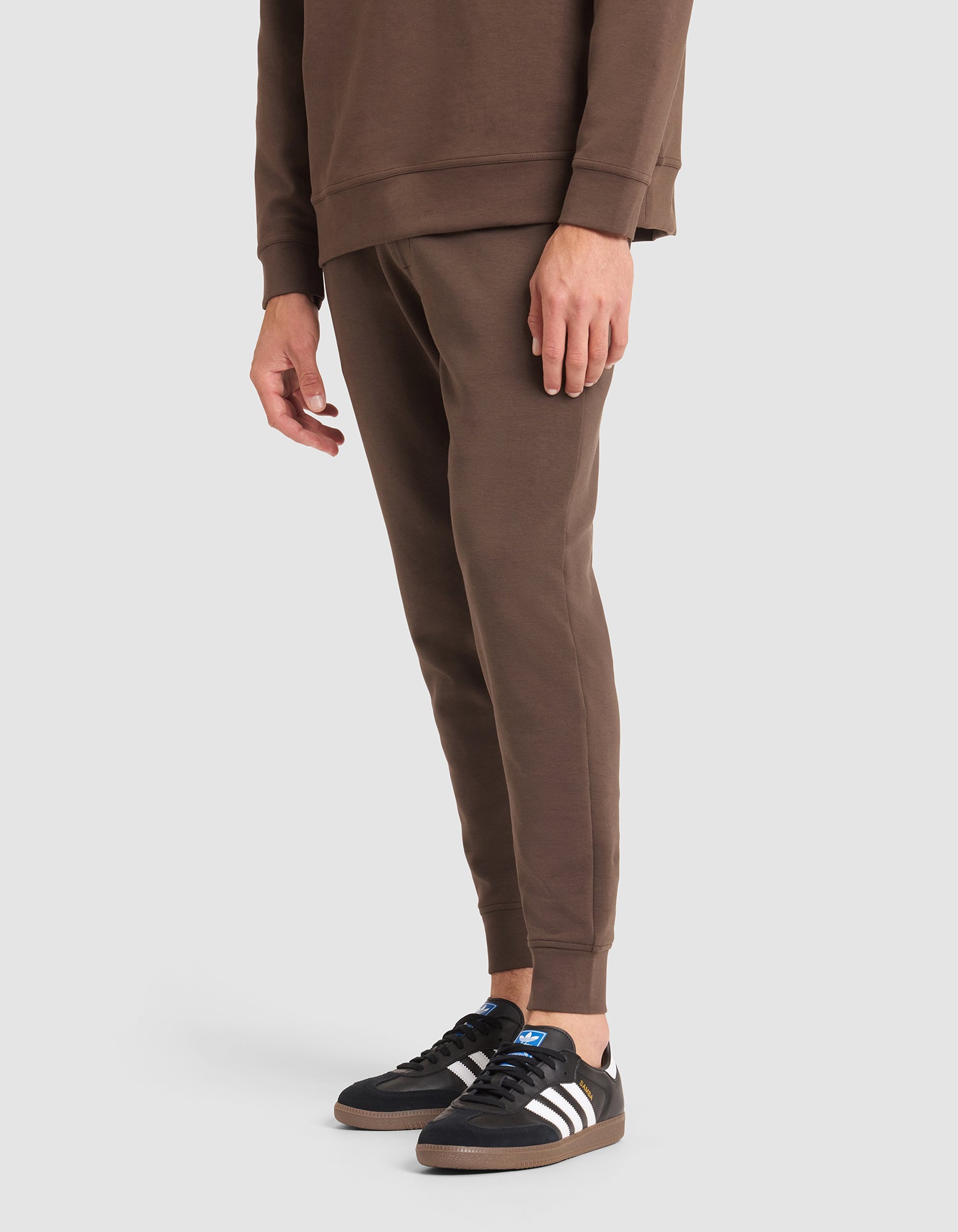 A person stands against a plain light background wearing a Cozy Earth Men's StretchTech Jogger set in brown and black Adidas sneakers with white stripes. 