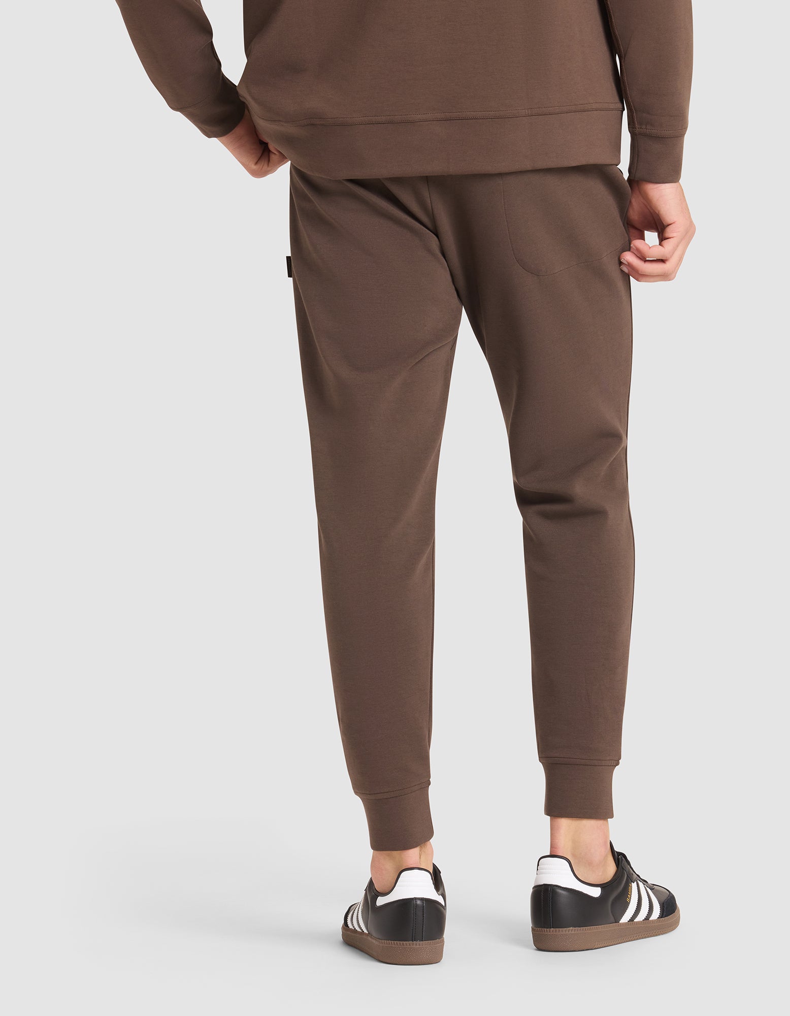 A person wearing Cozy Earth Men's StretchTech Jogger pants and a matching brown sweatshirt, paired with black Adidas sneakers with white stripes, is shown from the back against a plain light background. 