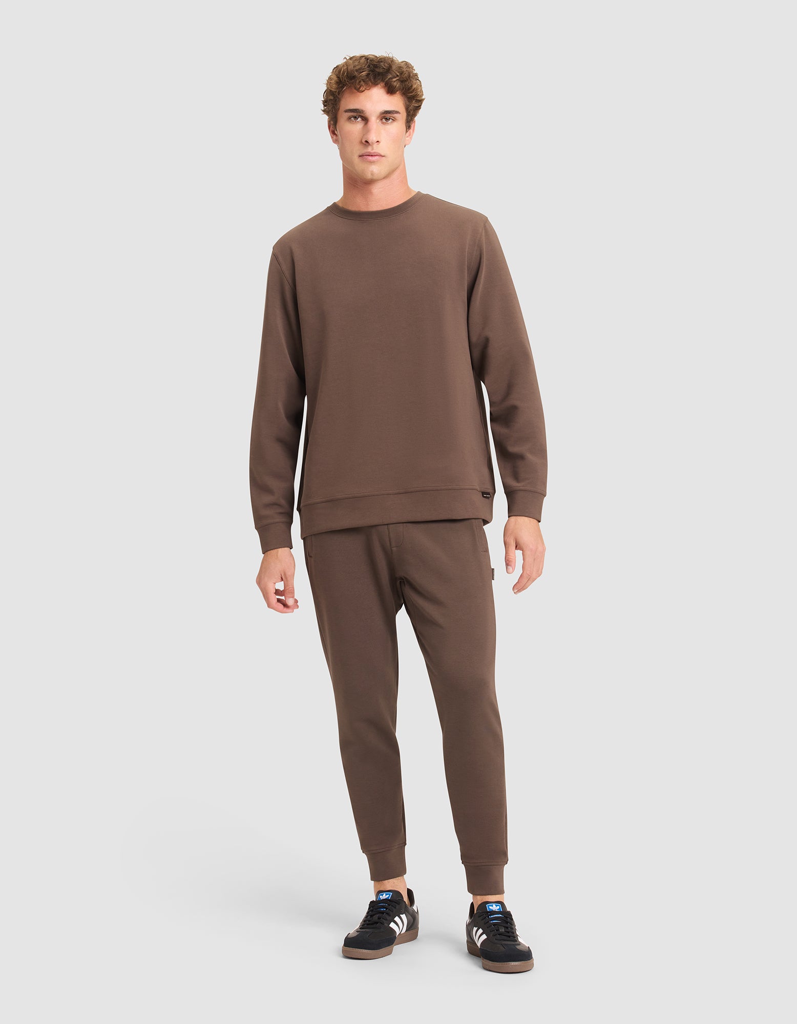 A person stands facing forward, wearing Cozy Earth's Men's StretchTech Jogger in brown with a matching sweatshirt and black sneakers featuring white soles and stripes, against a plain light gray background. 