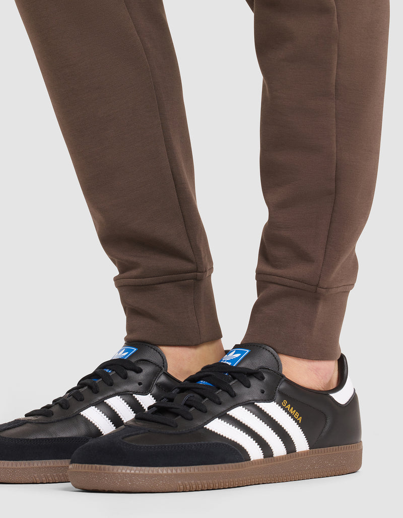 A person wearing Cozy Earth Men's StretchTech Jogger pants and black Adidas Samba sneakers with white stripes and gum soles stands on a light-colored surface.