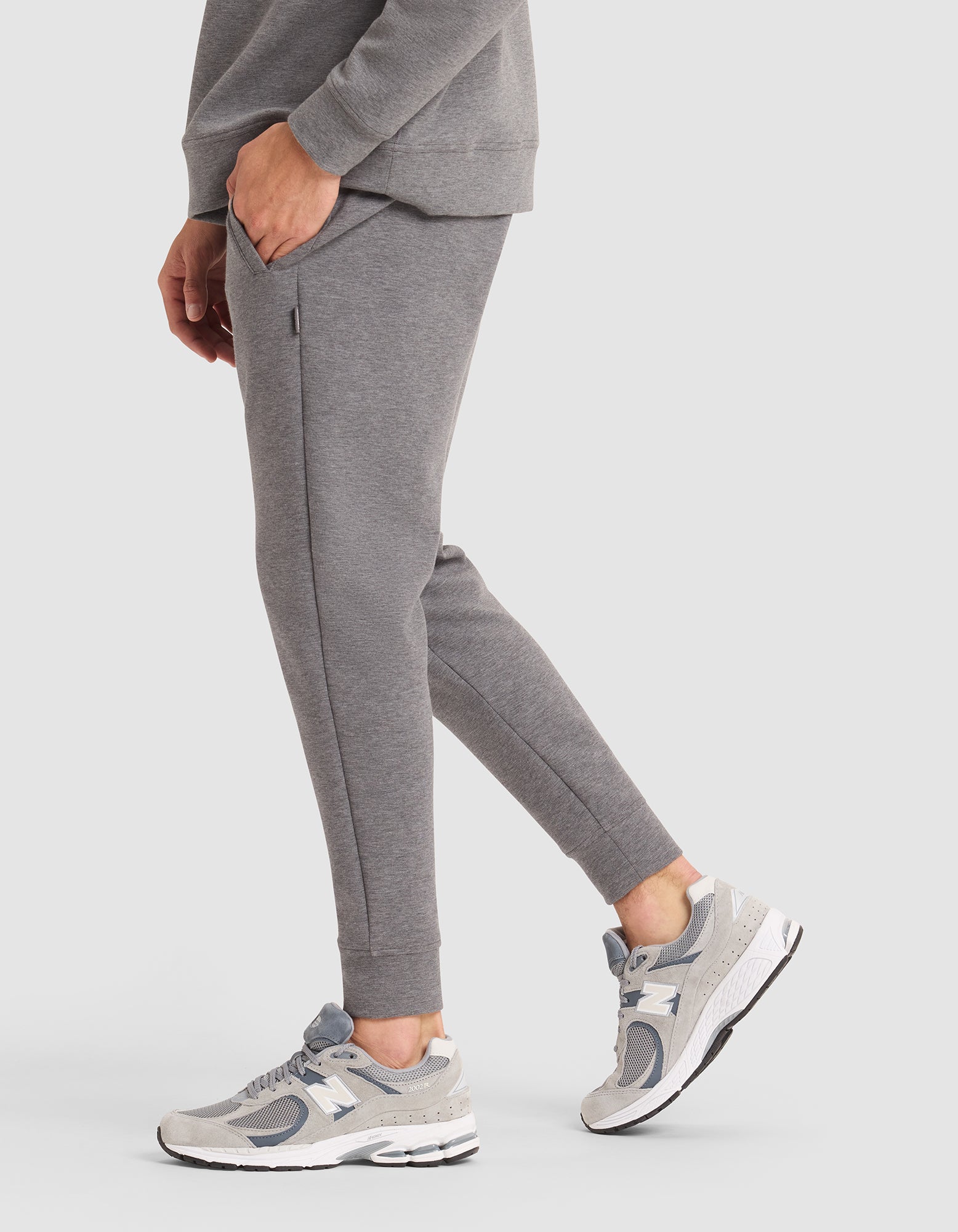 A person is shown from the shoulders down, wearing Cozy Earth's Men's StretchTech Jogger in grey with grey-and-white sneakers, standing on a white background with one hand in their pocket.  