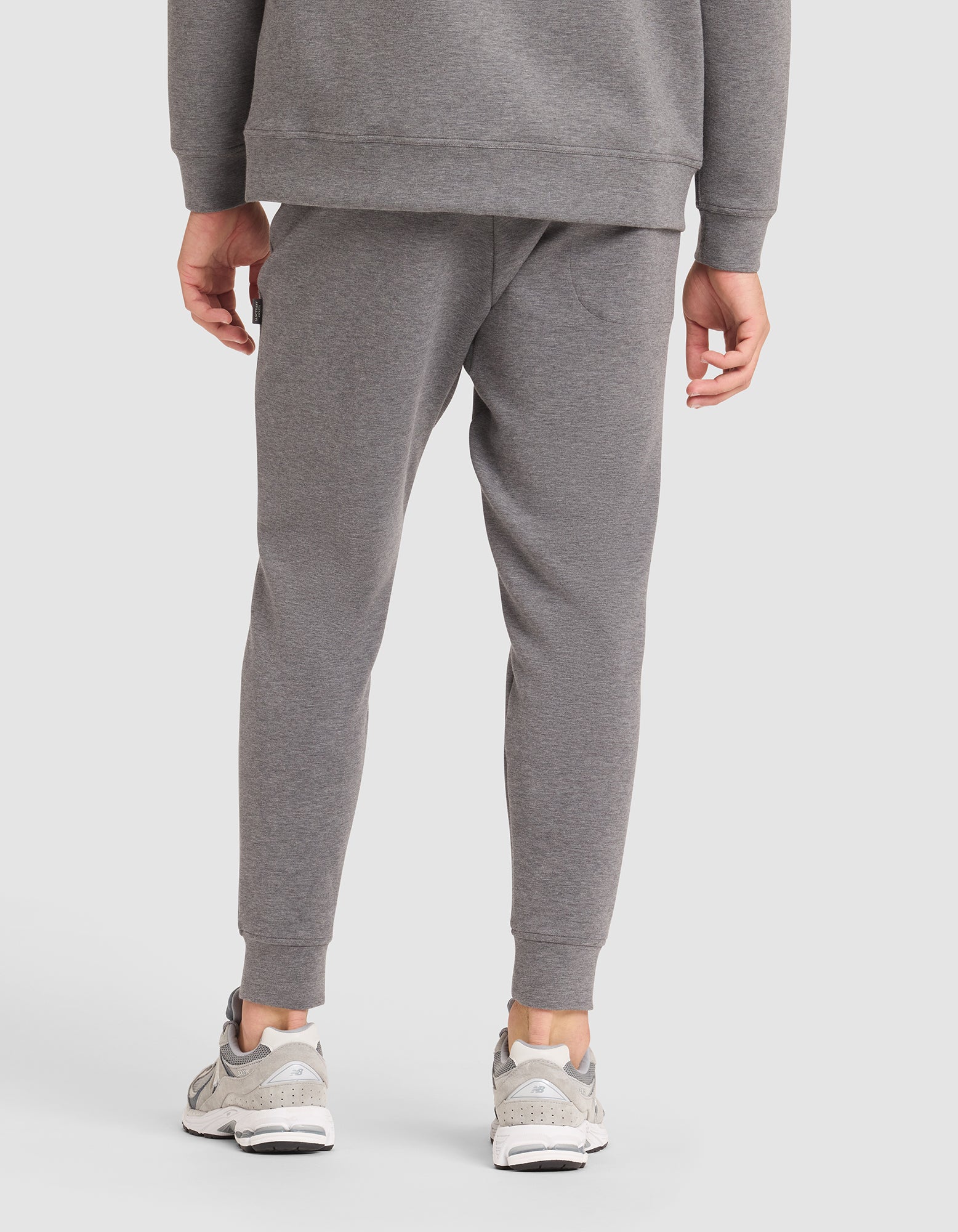 A person models the Cozy Earth Men's StretchTech Jogger in grey, paired with a matching sweatshirt and white sneakers, standing with their back to the camera against a plain white background. 